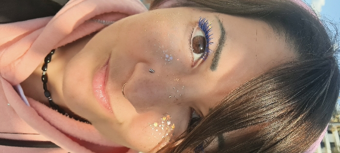 The image displays a close-up portrait of a female subject, positioned outdoors in Nahariyya, Israel. The subject has dark brown hair, parted on the left side, with some strands partially obscuring her right eye. Her left eye features bright blue mascara on both upper and lower lashes, and her irises are brown. Her skin has scattered glitter application; silvery-white iridescent glitter is visible on her left cheek and nose bridge, while golden-pink iridescent glitter is present on her right cheek. She has two nose piercings: a small, silver-colored stud on her left nostril and a gold-colored septum ring. Her lips are slightly parted in a subtle smile. She wears a light pink hooded sweatshirt. A black beaded choker-style necklace is visible around her neck. In the upper left background, a clear blue sky and a light-colored building structure with a railing are discernible, indicating an outdoor setting with natural daylight. A black strap, possibly from a bag or garment, is visible on the lower right edge of the frame.