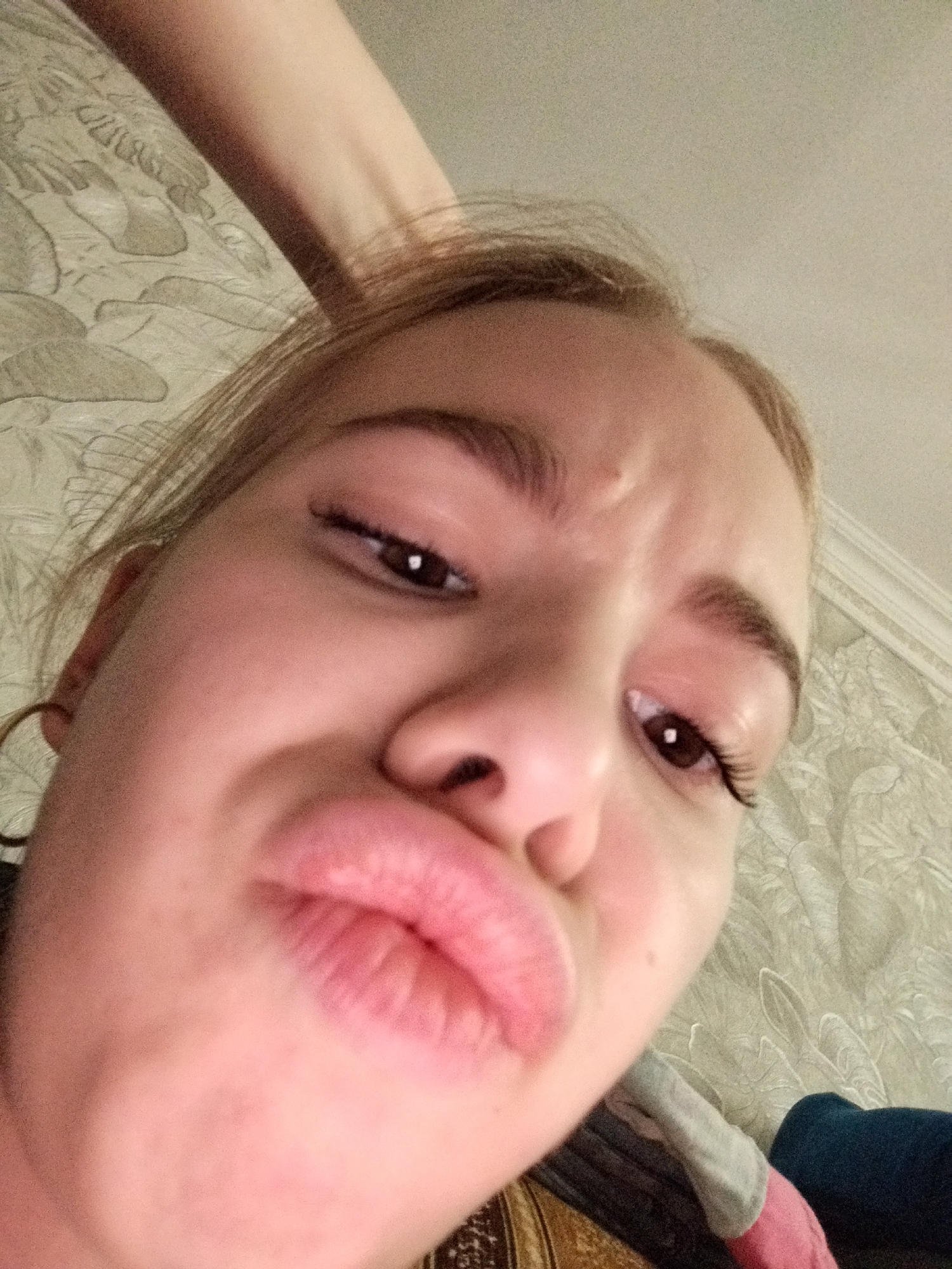 A young woman is captured in a low-angle, close-up selfie. She is making a puckered face, as if blowing a kiss, and her tongue is slightly visible. Her eyes are partially closed, and her eyebrows are furrowed. The background appears to be a patterned wallpaper. A portion of another person's arm is visible in the upper left of the frame, draped over what seems to be bedding or upholstery. The lighting is somewhat dim, casting a slight graininess over the image.