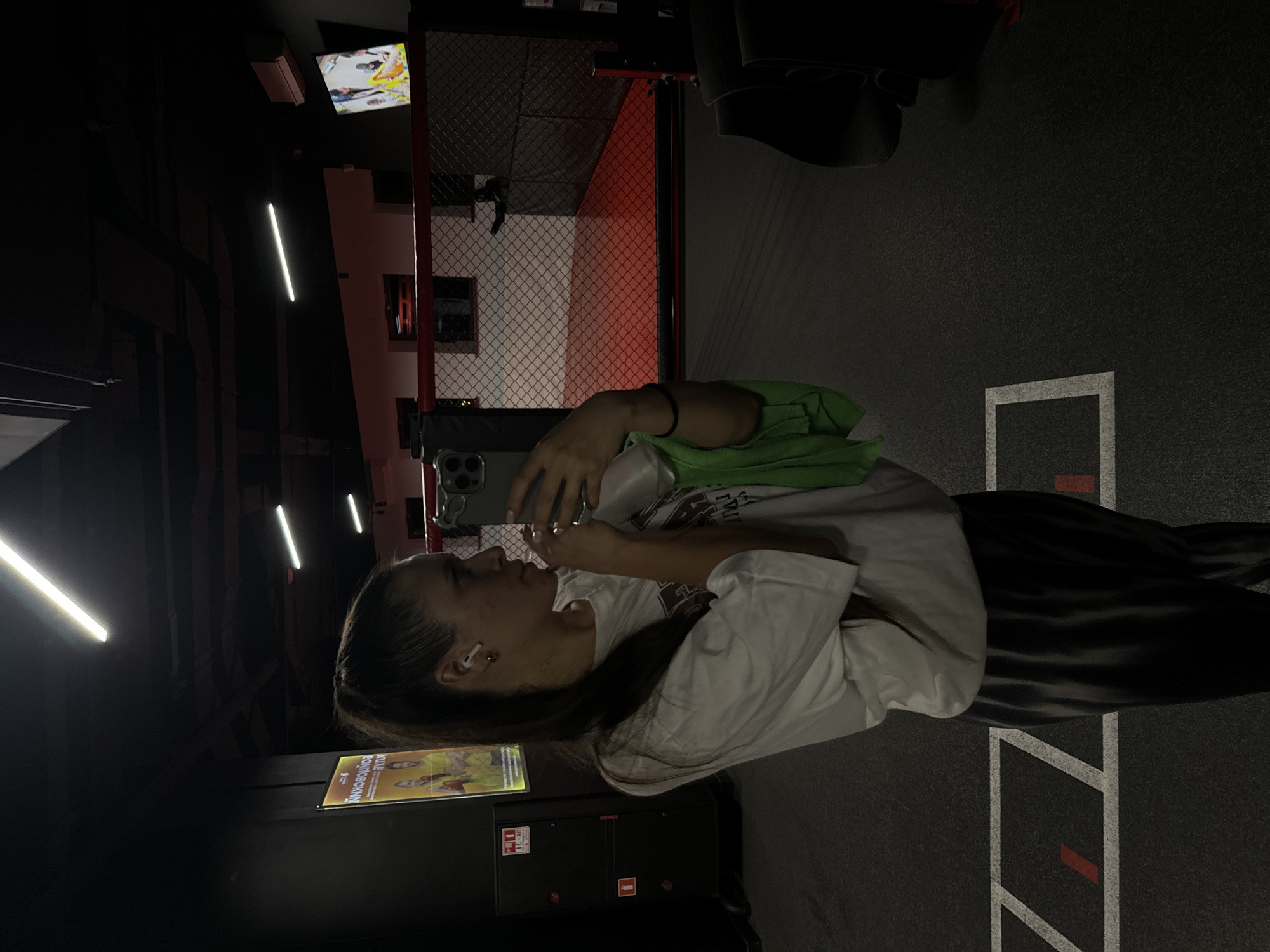 A woman stands in a dark gym in Vniissok, Russia, holding a smartphone. She is wearing a white t-shirt, dark pants, and has an earbud in her ear. A green bag is near her shoulder. The gym has a boxing ring with red accents, a television screen, and various workout equipment. Lights are visible on the ceiling. The floor is marked with white lines and red accents. A poster or sign is placed near the woman.