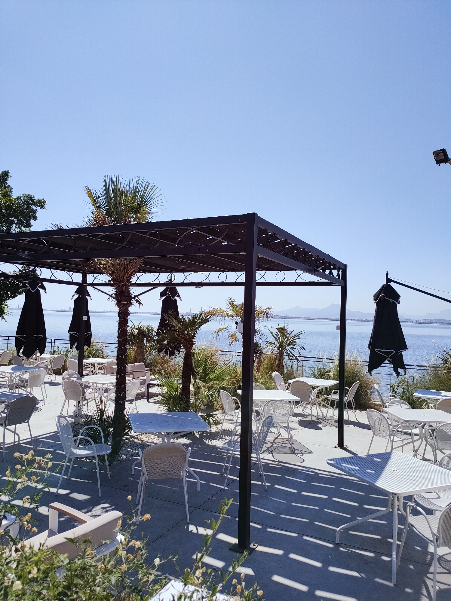 This image depicts an outdoor seating area for a restaurant or cafe, presumably located by a waterfront. The scene is characterized by numerous white tables and chairs arranged on a concrete patio. Several black umbrellas are positioned above some of the tables, providing shade. Palm trees and other foliage are visible, adding to the tropical or Mediterranean ambiance. In the background, a body of water, possibly a lake or sea, stretches towards a distant shoreline with mountains. The sky is clear and blue. The overall impression is one of a relaxed, outdoor dining environment. The image does not show any people or specific activities. The location is identified as Tunis, Tunisia.