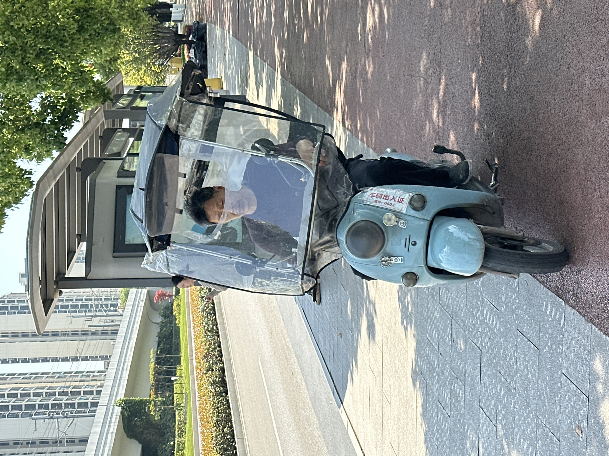 A man wearing a face mask is driving a light blue electric scooter with a transparent weather shield in Jiangjin, China. The scooter is parked on a sidewalk next to a street and a bus stop shelter. In the background, there are multi-story buildings, trees, and landscaped greenery. The sidewalk is made of light grey patterned paving stones, and the adjacent road has a white lane marking. Sunlight casts shadows of trees onto the sidewalk and road.