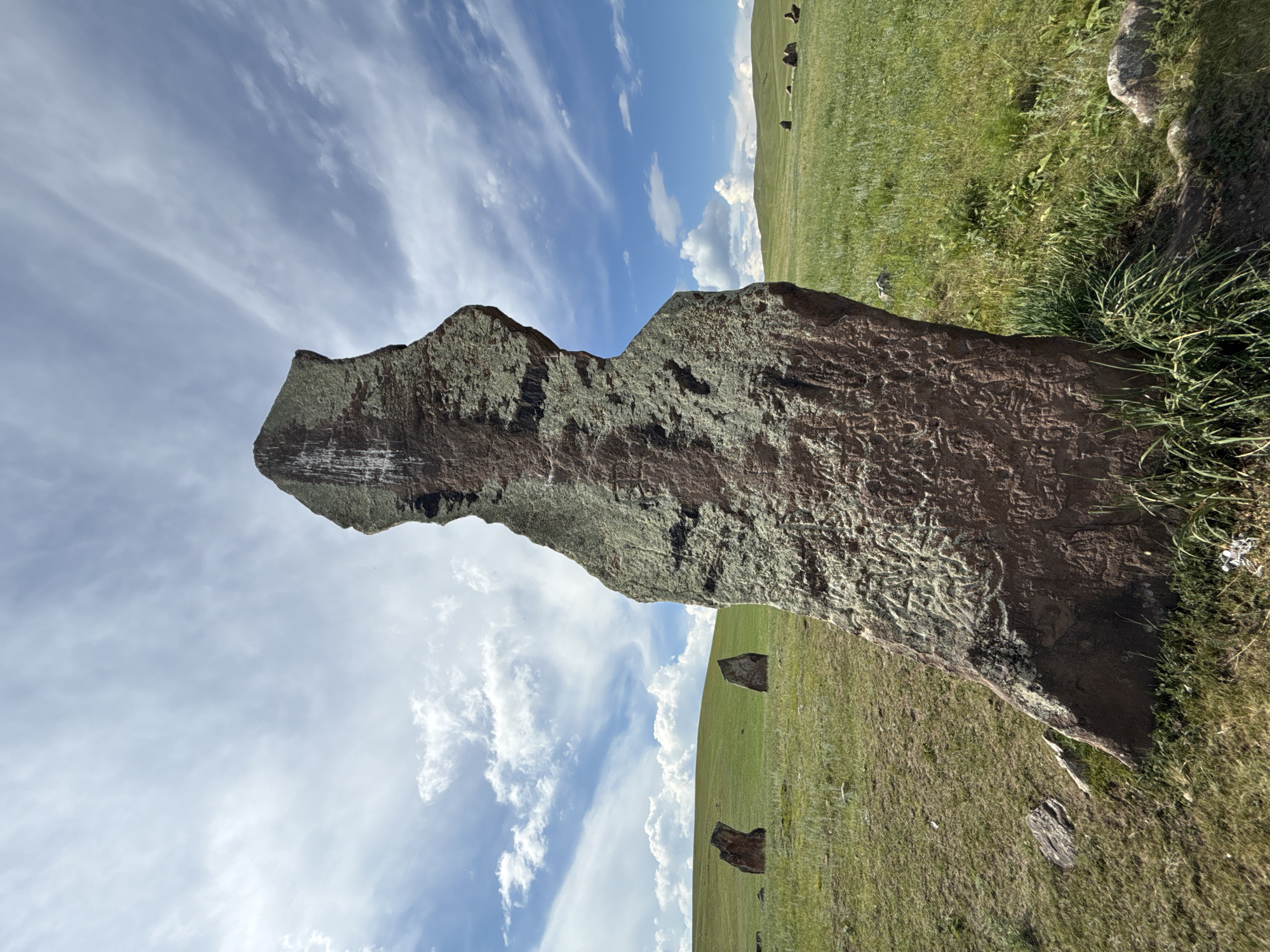This is a photograph taken in the natural landscape of Safronov, Russia. The image features a large, weathered stone in the foreground, partially covered in moss and lichen. The stone is intricately carved with patterns, suggesting it is an ancient artifact. Behind the stone, a rolling green meadow stretches into the distance, dotted with a few smaller, dark stones. The sky above is a vibrant blue, with streaks of white clouds that add depth and texture to the scene. The overall impression is one of natural beauty and historical significance. The lighting suggests it is daytime, with the sun casting shadows on the ground, and the clear sky indicates fair weather.