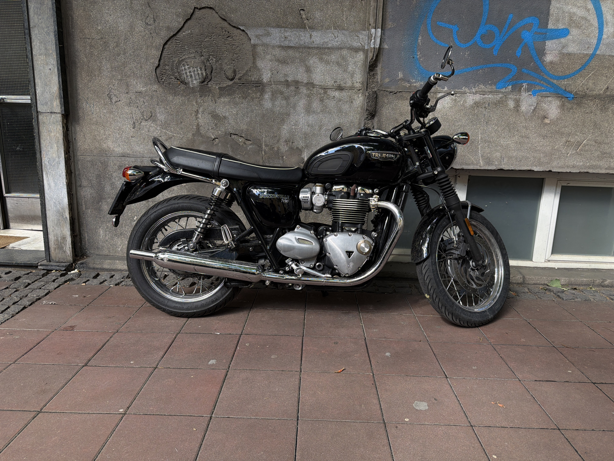 A black Triumph Bonneville T120 motorcycle is parked on a red-brick paved sidewalk in front of a gray concrete building in Belgrade, Serbia. The motorcycle is facing slightly to the right. Graffiti is visible on the concrete wall behind the motorcycle. A doorway is to the left, and a window is to the right of the motorcycle.