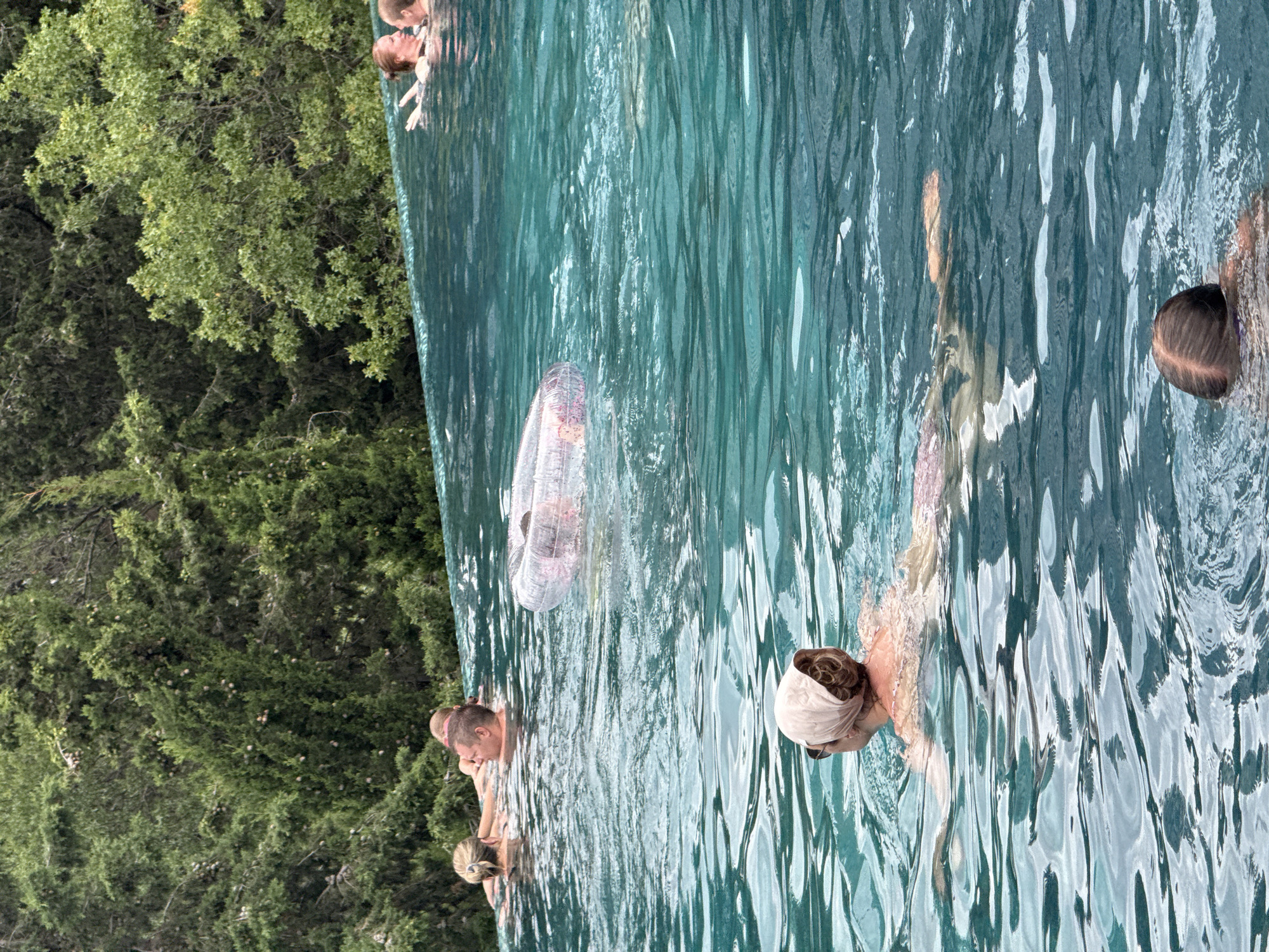 In the upper portion of the image, a group of people are visible in an infinity pool with a lush green forested backdrop. In the foreground of this section, two women and a man are partially submerged in the water. The man is looking towards the left, while one woman is looking at him and the other is facing away from the camera. Further back, another woman is visible. To the right of this group, two more women are interacting in the pool. A transparent inflatable ring with a pink pattern floats in the middle of the pool.
The lower portion of the image focuses on a person swimming in the foreground of the pool, wearing a light-colored headscarf and sunglasses. The person is swimming away from the camera, with their body partially submerged. In the far lower right corner, another person's head is visible, also swimming away from the camera. The water is a vibrant turquoise color, and ripples from the swimmers are visible on its surface. The overall environment suggests a recreational setting, likely a hotel or private residence with a pool overlooking a natural landscape.