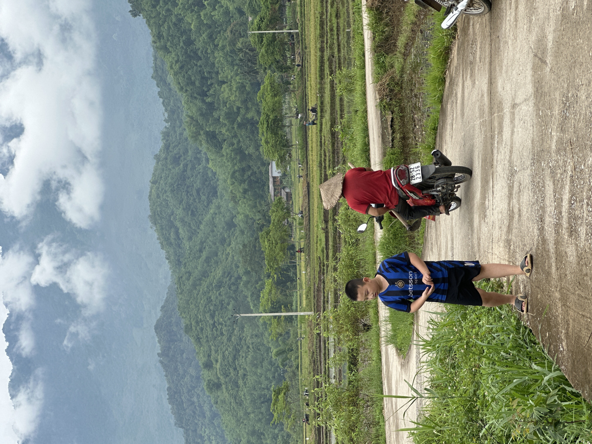 In Mai Chau, Vietnam, a young boy in a blue and black striped soccer jersey and black shorts stands on a dirt path. To his right, a person wearing a red shirt and a conical hat rides a red motorcycle. The path leads towards a verdant landscape of rice paddies with terraced fields, where several people are working. In the background, rolling green hills rise up, partially obscured by a cloudy sky.