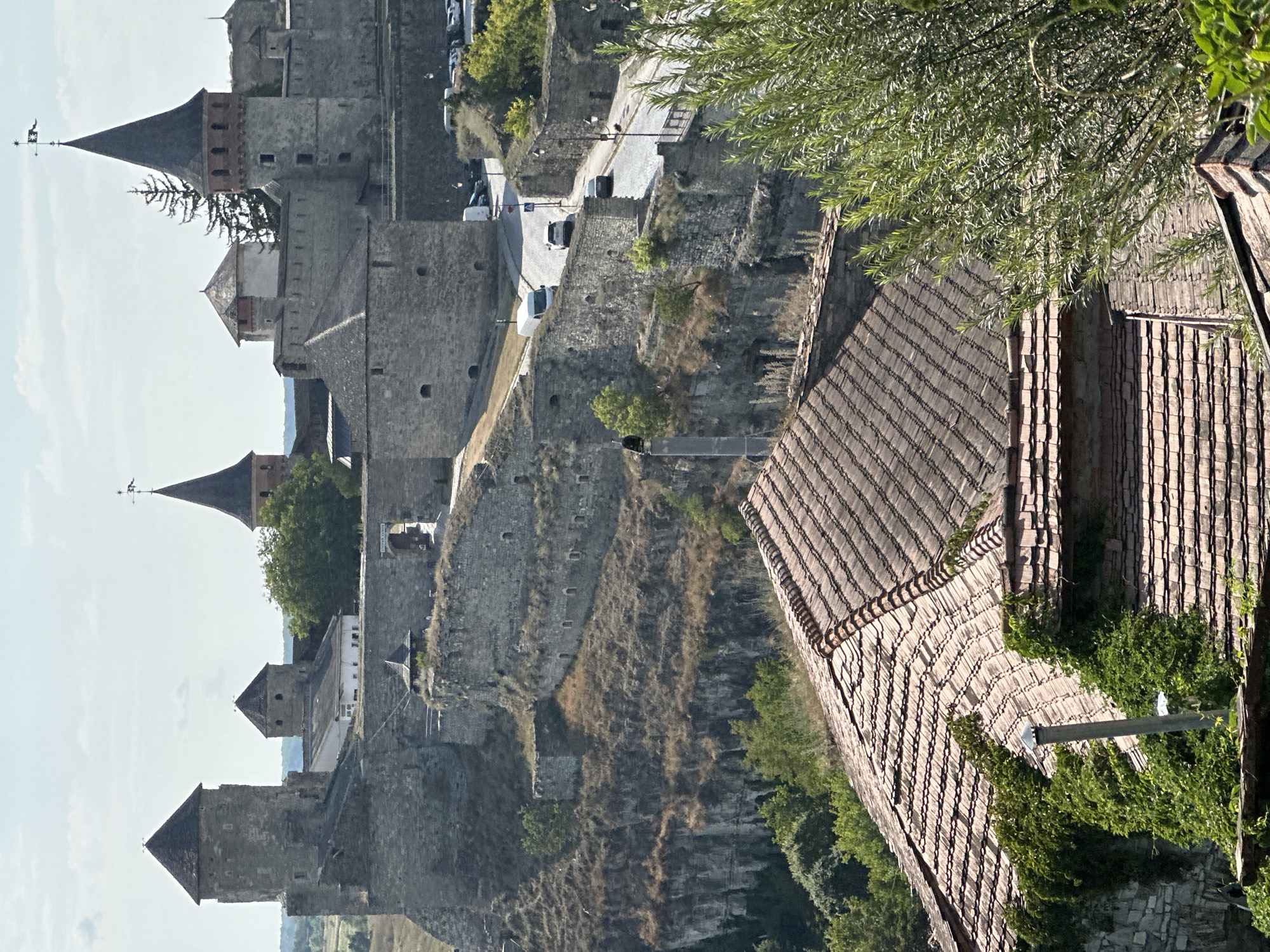 The image depicts a panoramic view of the Kamianets-Podilskyi Castle in Kamianets-Podilskyi, Ukraine, under a clear sky. The ancient stone fortress, a prominent historical landmark, occupies the upper portion of the frame, characterized by its imposing walls, turrets, and conical roofs. A road winds its way through the castle grounds, with a few vehicles, including cars and vans, visible. The castle is perched on a rocky, elevated terrain, with a steep cliff face visible on the left, covered in dry grass and sparse vegetation. In the foreground, the tiled roofs of several buildings, partially obscured by greenery, suggest the presence of a town or settlement adjacent to the historical site. The overall scene conveys a sense of historical grandeur and the integration of a medieval fortress with its surrounding environment.
