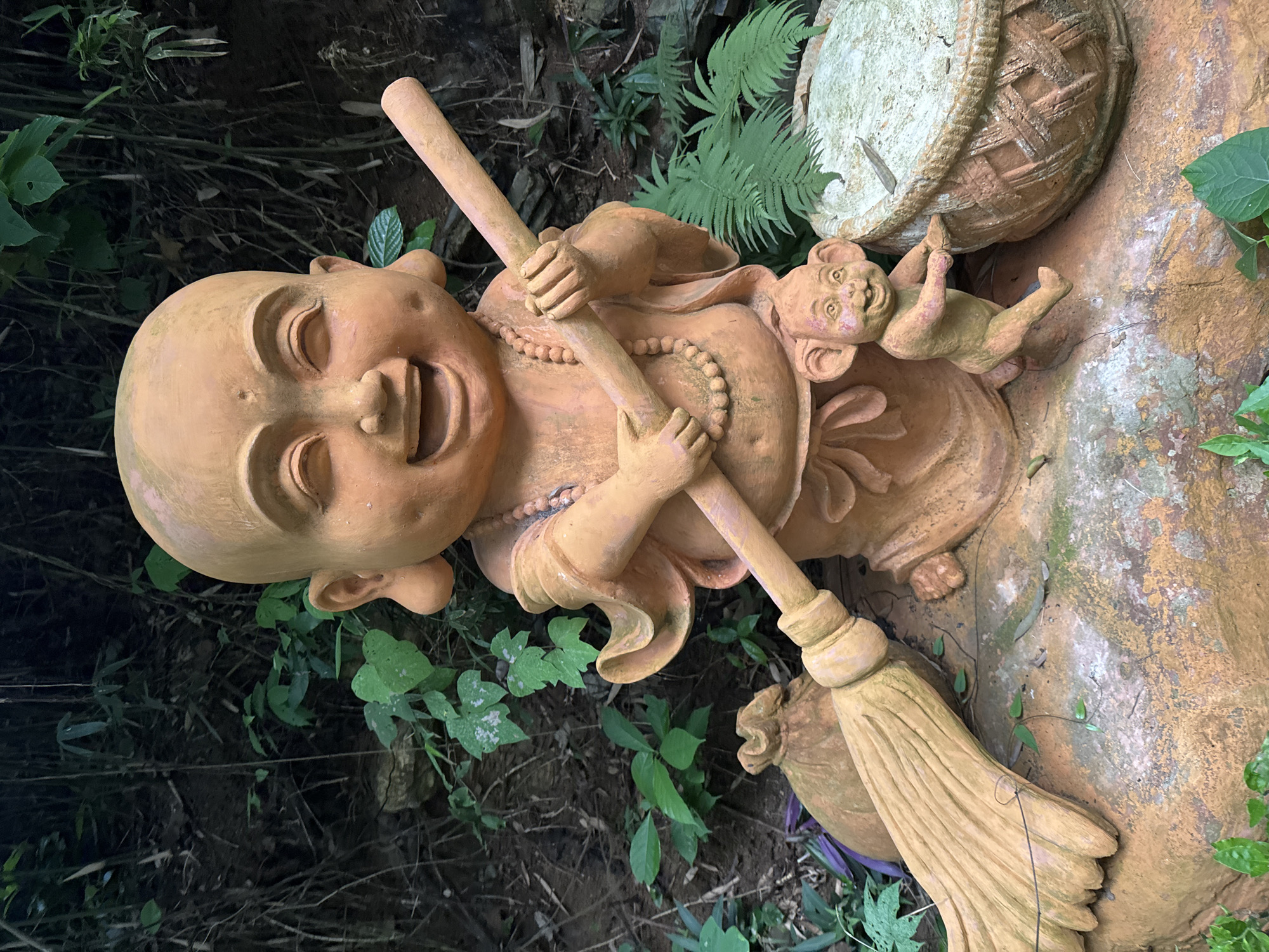 A full shot shows a terracotta statue of a laughing child monk holding a broom in a courtyard. The monk statue is positioned in the center of the frame, kneeling on a large rock and smiling broadly with his eyes closed. He wears a beaded necklace and is holding a long, wooden broom with his arms crossed. To the left of the monk, a small, brown sack with a rope tie rests on the rock. To the right of the monk, a small, mischievous-looking monkey statue stands with its hands on a woven basket. The background consists of dense green foliage and dark, rocky terrain. The overall scene is vibrant and playful, suggesting a garden or outdoor decorative setting in the Tây Hoa Lư Ward of Vietnam.