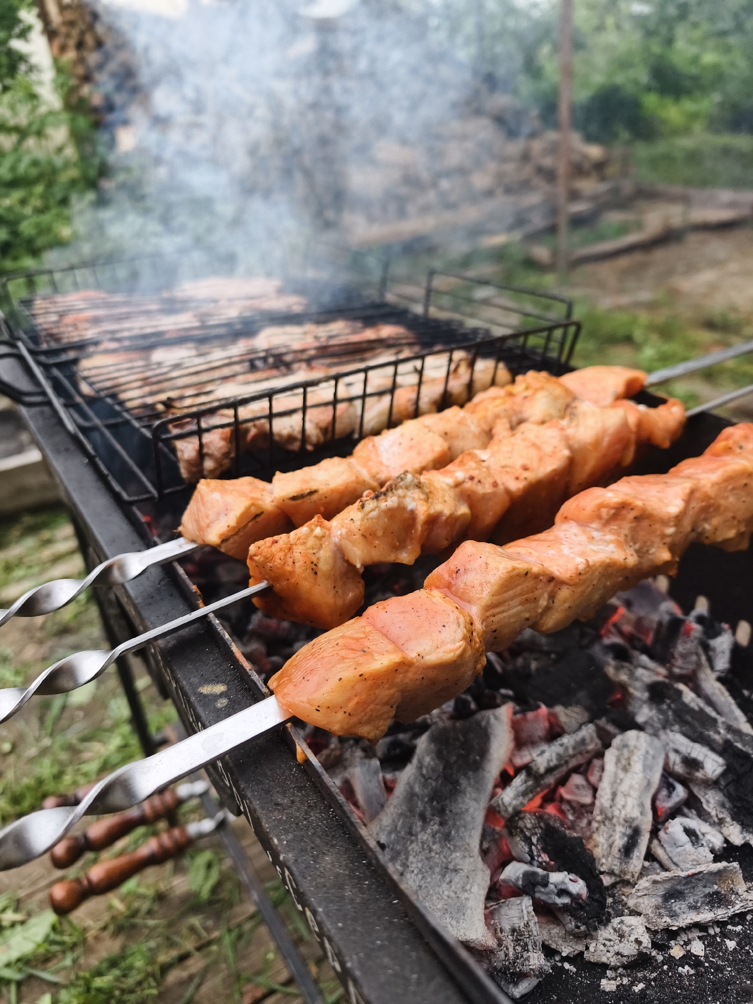 The image displays an outdoor cooking scene in Prikamskii, Russia. In the immediate foreground, three metal skewers (shampurs) are positioned over a black metal brazier (mangal). Each skewer holds multiple cubed pieces of reddish-orange, marinated meat, likely salmon or pork, which exhibit scattered dark specks of seasoning. Beneath the skewers, the brazier contains hot, glowing red coals interspersed with white and grey ash. To the left of the skewered meat, a black metal grill grate or grilling basket is visible, holding additional pieces of meat also in the process of cooking. A substantial plume of light grey smoke rises from the cooking area. The background is out of focus but shows various green elements consistent with foliage, including trees and bushes, along with indistinct rustic structures, indicating an outdoor, possibly rural or semi-rural environment. The ground surrounding the brazier appears to be a mix of dirt and sparse green grass.