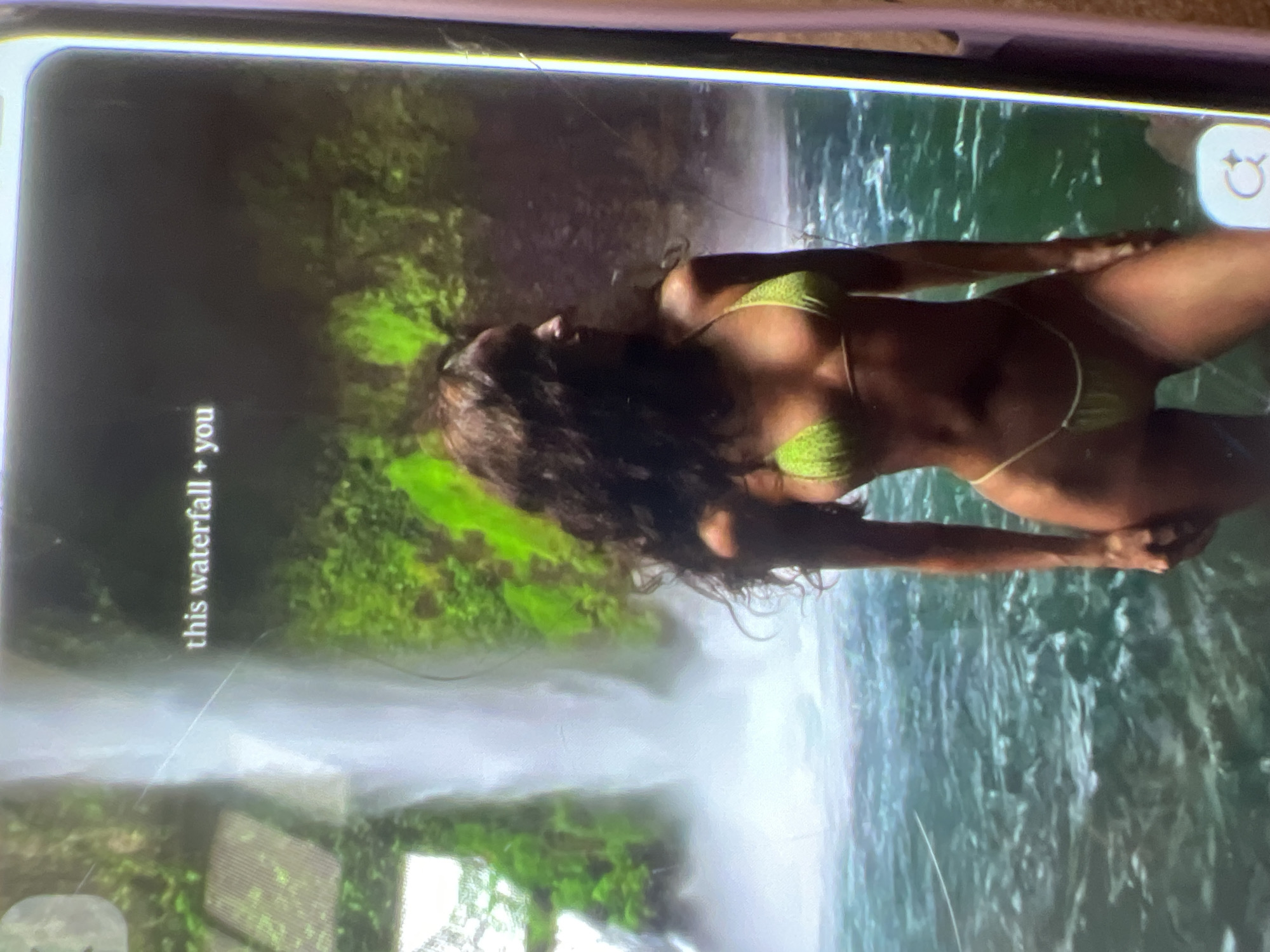 A young woman in a green bikini stands in front of a waterfall. The woman has dark, curly hair and is facing to the right. The waterfall is surrounded by lush green foliage. The water in the foreground is a deep blue-green. There is text overlaid on the image that reads "this waterfall + you." The image appears to be a screenshot from a mobile device. There is no indication of Kara-Dıykan, Kyrgyzstan in the image.
