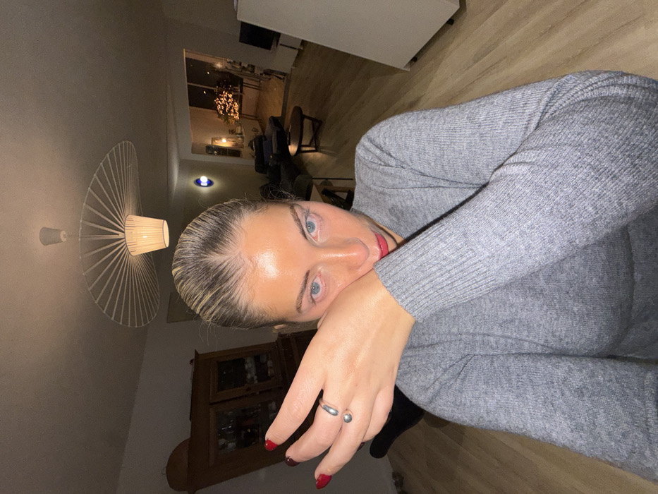 A woman with blonde hair and blue eyes poses for a selfie. She wears a gray sweater and has on red lipstick. Two silver rings adorn her right hand. In the background, a modern lamp hangs on the wall. A cabinet with glass doors and what appears to be a doorway leading outside are also visible. The photo is taken at an odd angle with the camera facing up toward the subject.