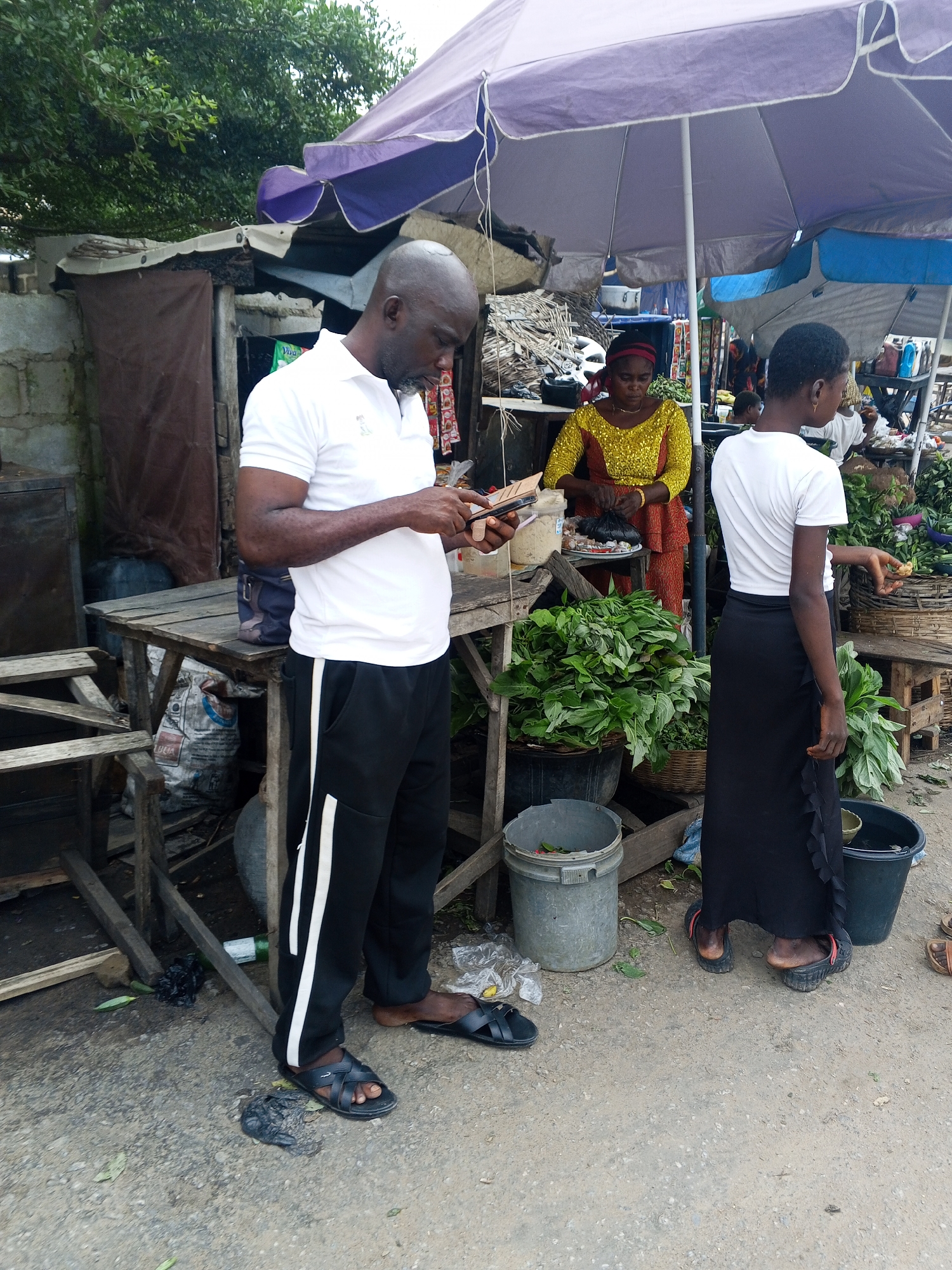 In Zango-ndaji, Nigeria, a man in a white polo shirt and black pants with white stripes, wearing black sandals, stands on a dirt ground, looking at his phone. Behind him, under a purple umbrella, a woman in a yellow and red patterned dress is visible, as well as another young woman in a white t-shirt and black skirt. The environment appears to be a market, with various stalls, baskets of vegetables, and buckets present. The ground is uneven and dusty.
