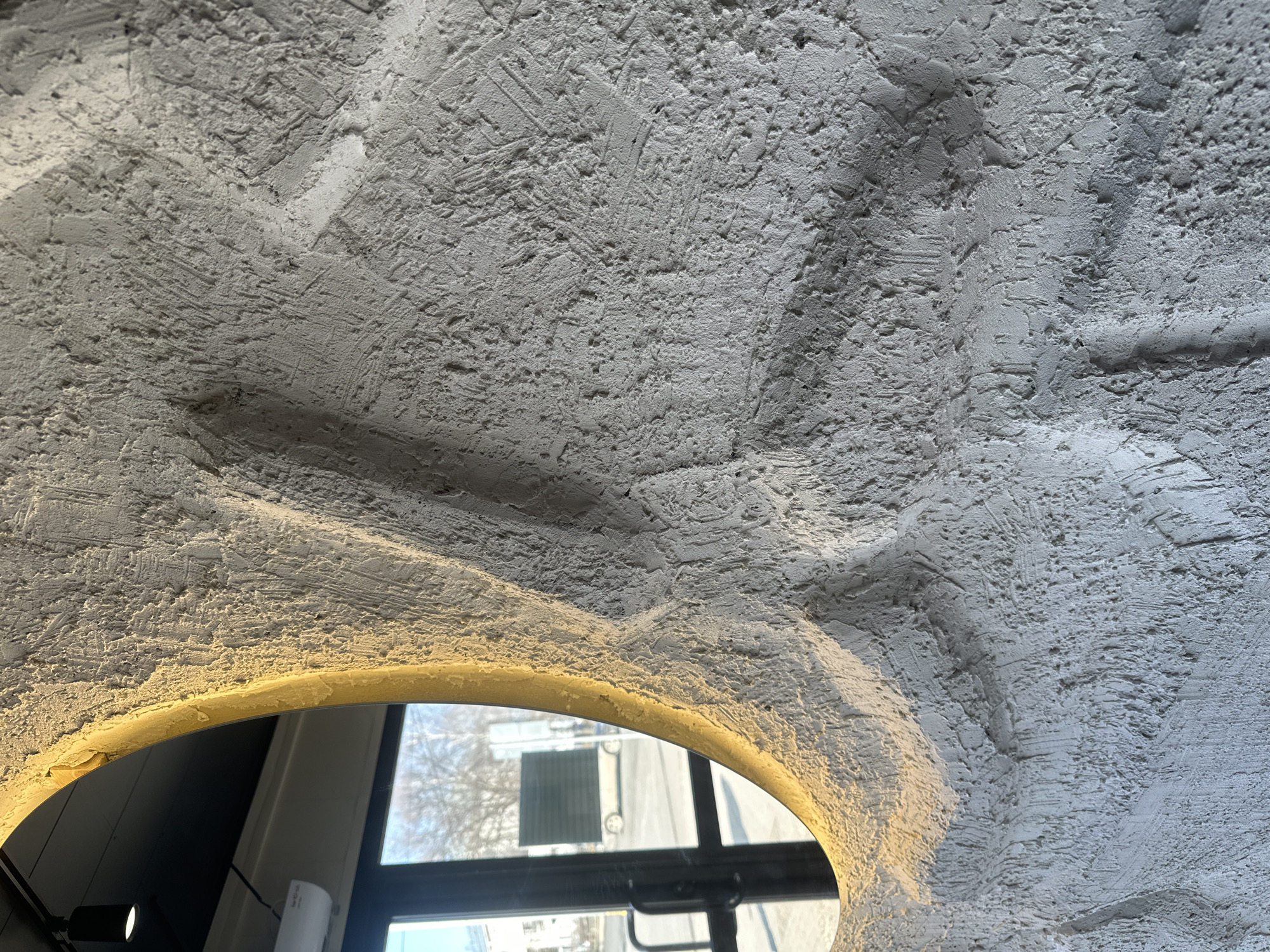 A close-up, slightly angled view focuses on a textured, light-gray or white ceiling or wall with an arched opening revealing a window and exterior scene. The wall surface is rough, with visible trowel marks and an uneven, grainy texture.
The arched opening is illuminated with a warm, yellow light around its perimeter. Through the opening, a window is visible. Outside the window, a building with a symmetrical facade and a grid of windows can be seen. Bare trees without leaves stand against a light sky.
A portion of a black light fixture is visible in the lower left corner of the frame, and a small white device is partially visible to the left of the window.
Without additional information or context, it is not possible to definitively determine the location in Chaikovsky, Russia.