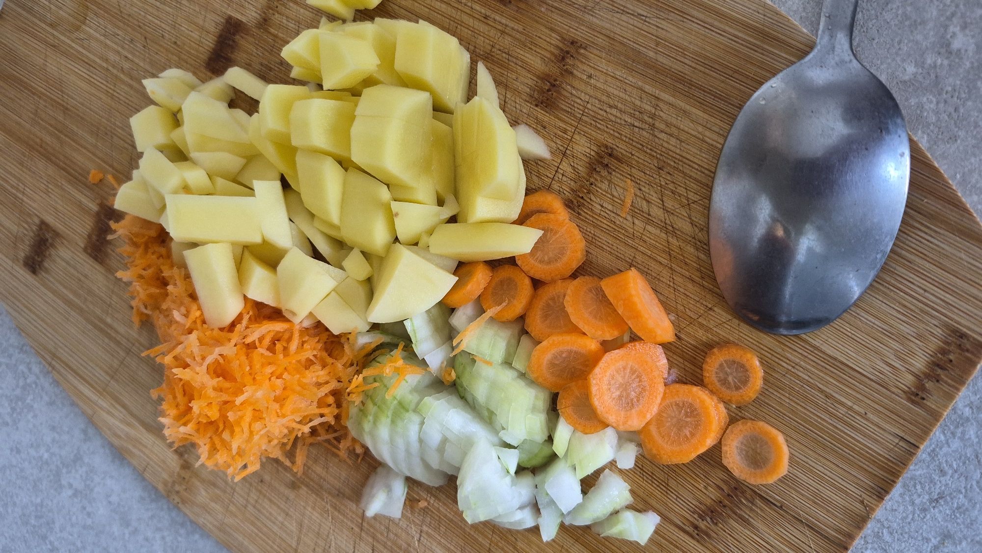 In Krauja, Latvia, a close-up overhead shot captures chopped vegetables on a wooden cutting board. The scene is devoid of people. From left to right, a pile of finely grated carrots is visible, followed by chopped white onions. Next to the onions are sliced carrots in rounds and some angled pieces. To the right of the vegetables, diced yellow potatoes fill a significant portion of the board. In the lower right corner, a metal spoon rests on the cutting board. The lighting is neutral, highlighting the textures and colors of the ingredients.