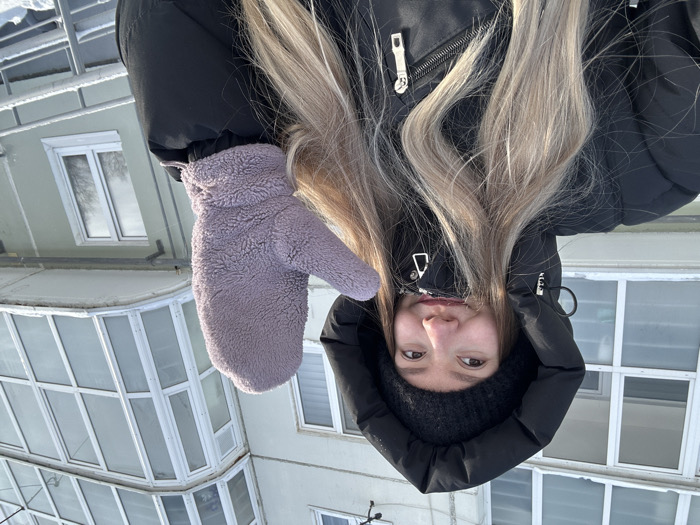 A young adult female is captured outdoors on a bright, sunny winter day in Perm, Russia. She stands in the foreground, smiling faintly and looking towards the right of the frame. Her long blonde hair is partially visible beneath a black ribbed beanie and the raised hood of a black puffer jacket. Her right hand is elevated, prominently displaying a light purple, fluffy mitten. The background features a multi-story, light grey residential building with multiple white-framed windows and an enclosed, angular balcony on an upper floor. The ground in the foreground, building ledges, and a metal railing in the lower right are all covered in snow, consistent with a clear, cold winter environment.