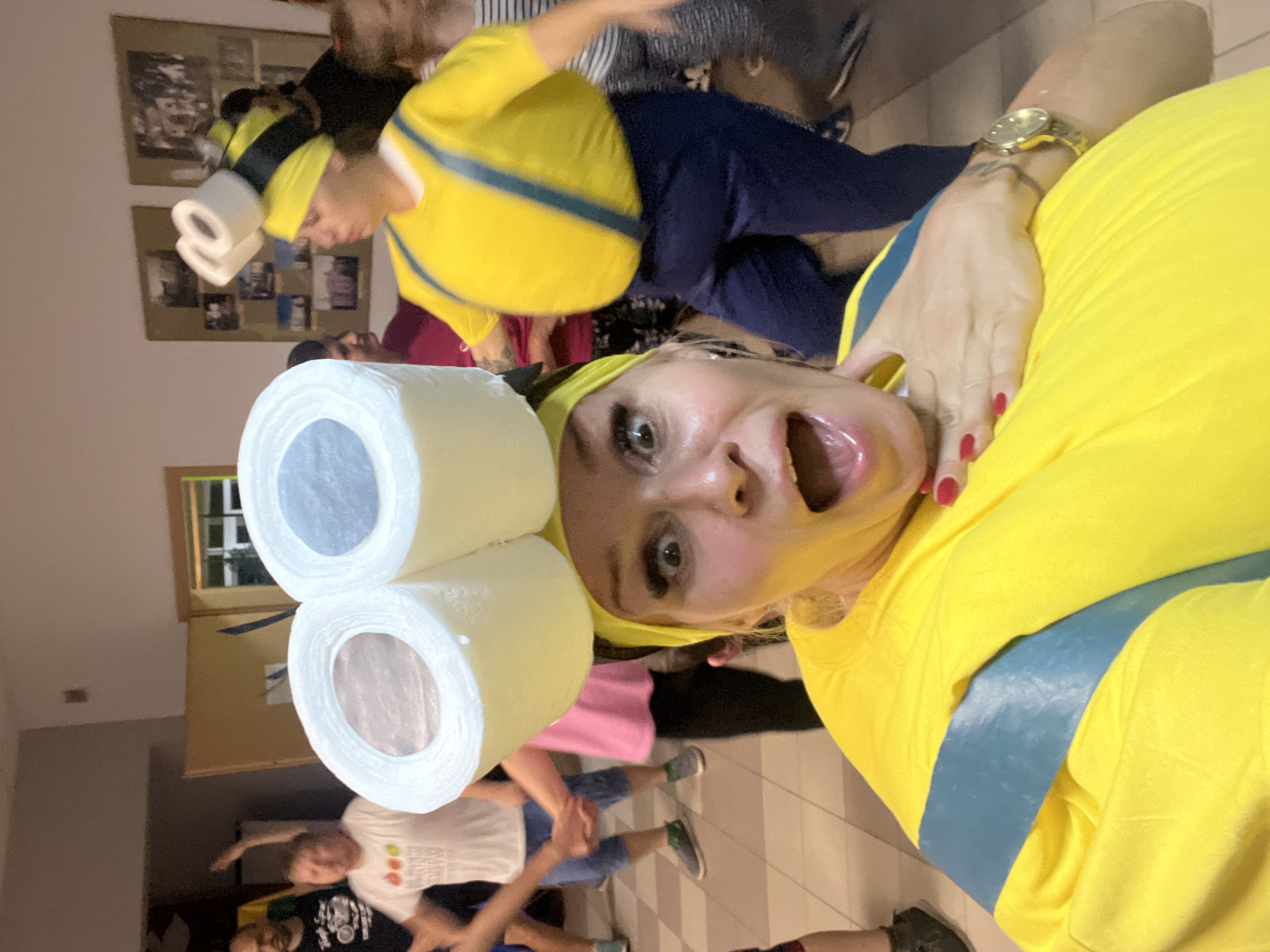In Hodyszewo, Poland, a woman is dressed in a yellow minion costume with blue overalls and a yellow headband, sporting two toilet paper rolls on her head as a hat. She has a shocked expression with her mouth open. Another person in the background also appears to be in a minion costume. The setting is indoors, possibly a community hall or event space, with a tiled floor and framed pictures on the wall. Other people are visible in the background, some dancing.