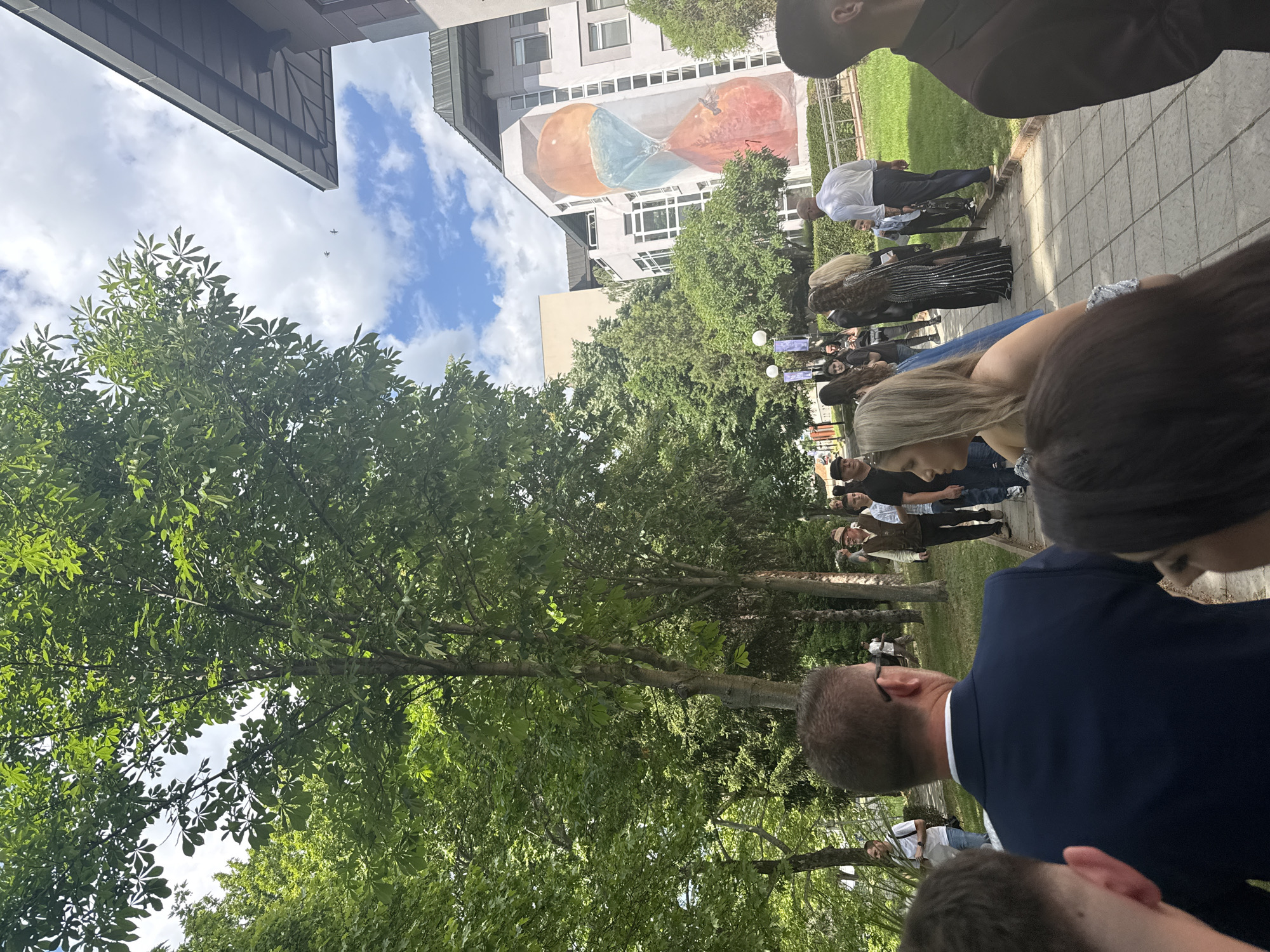 The image shows a gathering of people outdoors, likely an event, in the city of Blagoevgrad, Bulgaria. A tall building with a mural depicting an hourglass is visible in the background, indicating the location. The foreground is partially obscured by a large tree with dense green foliage. The people are dressed in a mix of formal and casual attire. Some are standing and others appear to be interacting with each other. The sky is partly cloudy.