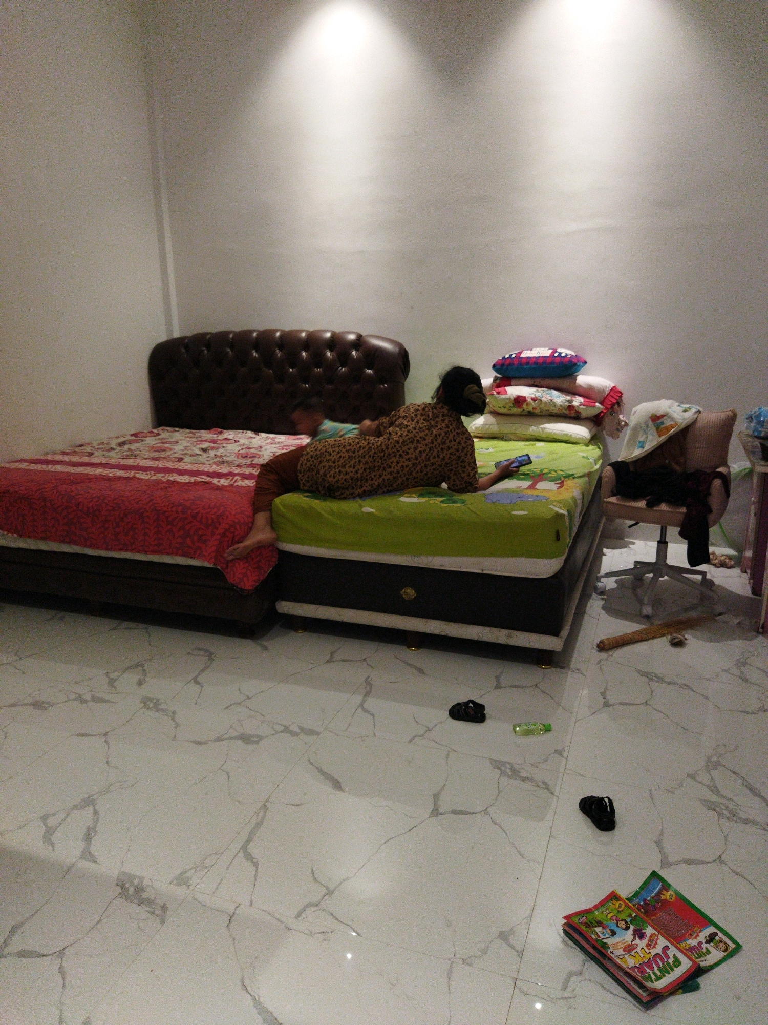 The image depicts a bedroom interior in what is identified as the city of Banjar Agung, Indonesia. The room features two beds side-by-side. The bed on the left is made with a red and white patterned comforter and a dark brown tufted headboard. Adjacent to it, the bed on the right is covered with a green sheet and appears to have a stack of pillows at the head. A person, with dark hair and wearing a patterned shirt and brown pants, is lying on the right bed, holding a mobile phone. The floor is tiled with a white marble pattern. To the right of the beds, a swivel chair with a brown cushioned seat is positioned, with clothes draped over it and a broom lying on the floor nearby. Several items, including black flip-flops and a small bottle, are scattered on the floor. Two colorful, comic-like books are also stacked on the floor in the foreground. The walls are painted white, and two spotlights are visible on the ceiling, casting light onto the wall.