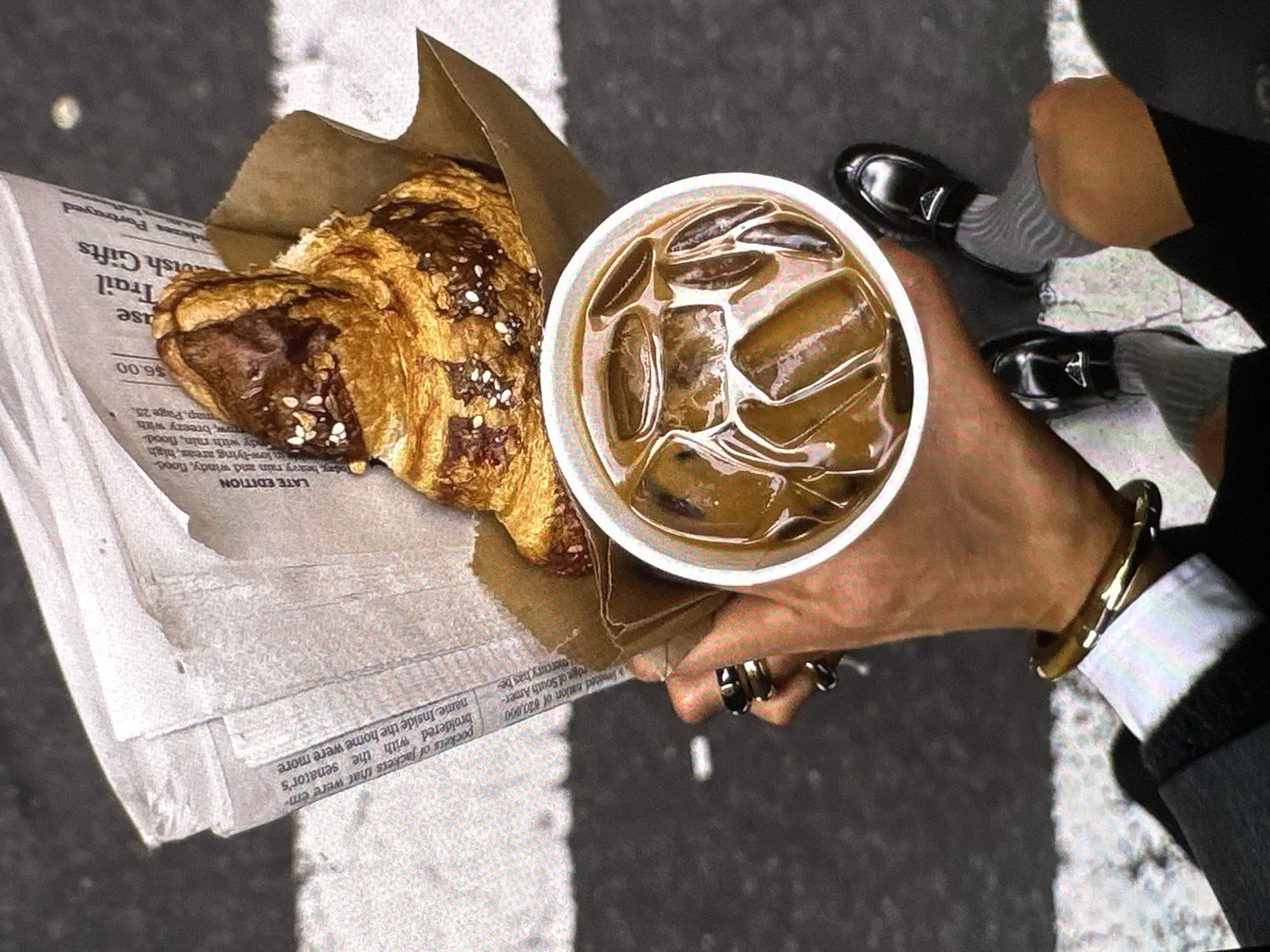 The image shows a person holding a newspaper, a croissant in a brown paper bag, and a cup of iced coffee. The person is wearing a black suit, white shirt, gray socks, and black loafers. They are also wearing rings on their fingers and a bracelet on their wrist. The newspaper has the words "LATE EDITION" printed on it. There is no indication of the city Yerevan, Armenia.