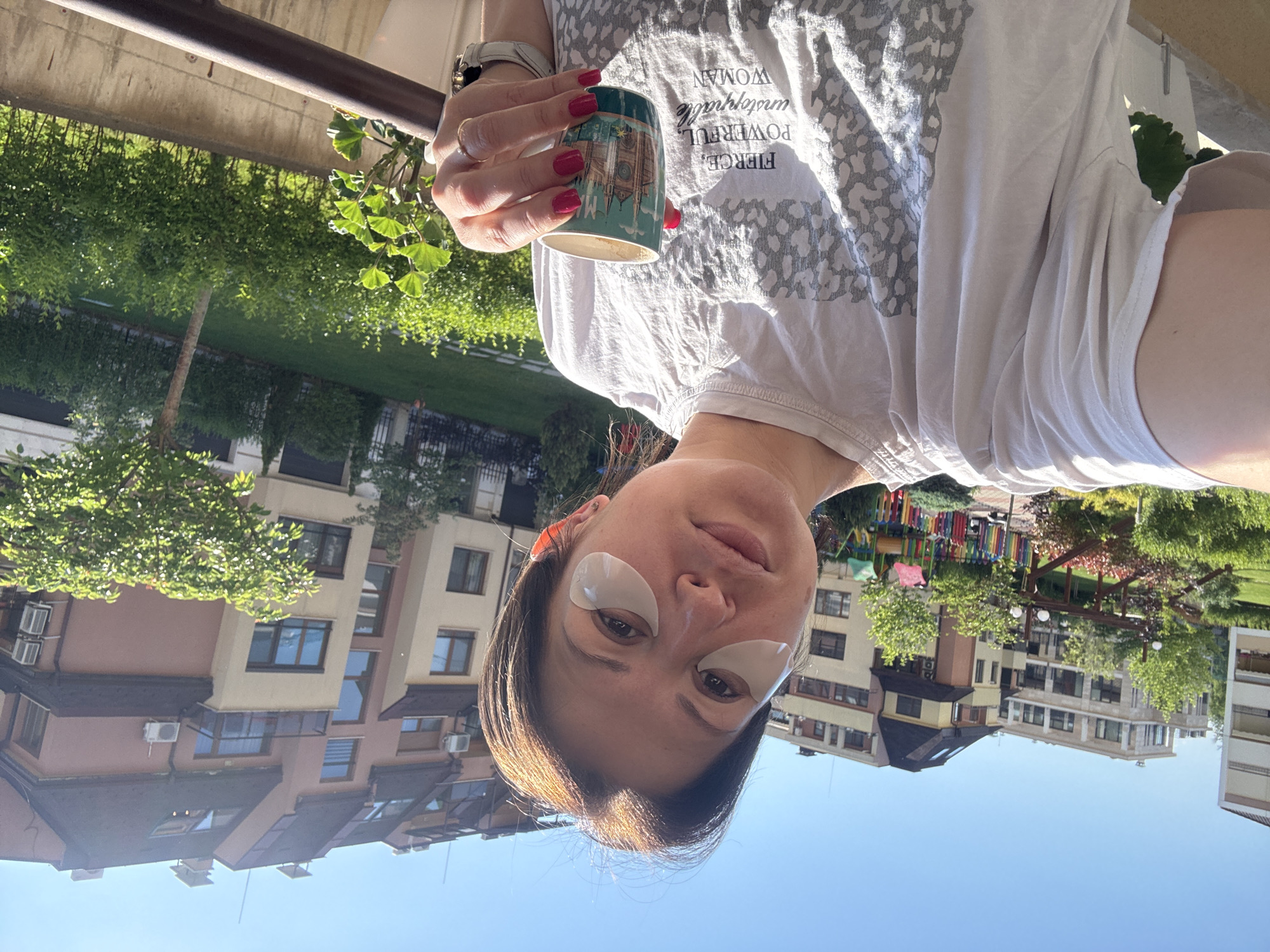 A woman with undereye patches is holding a cup, outdoors. The cup is light blue and has what appears to be a depiction of a cathedral. The woman's nails are painted red. She is wearing a light-colored t-shirt with grey animal print and the words "FIERCE. POWERFUL. unstoppable WOMAN" written on it. The background features a building complex with a playground, foliage, and sky. Based on architectural style and landscaping, this is likely a residential complex in Sofia, Bulgaria.