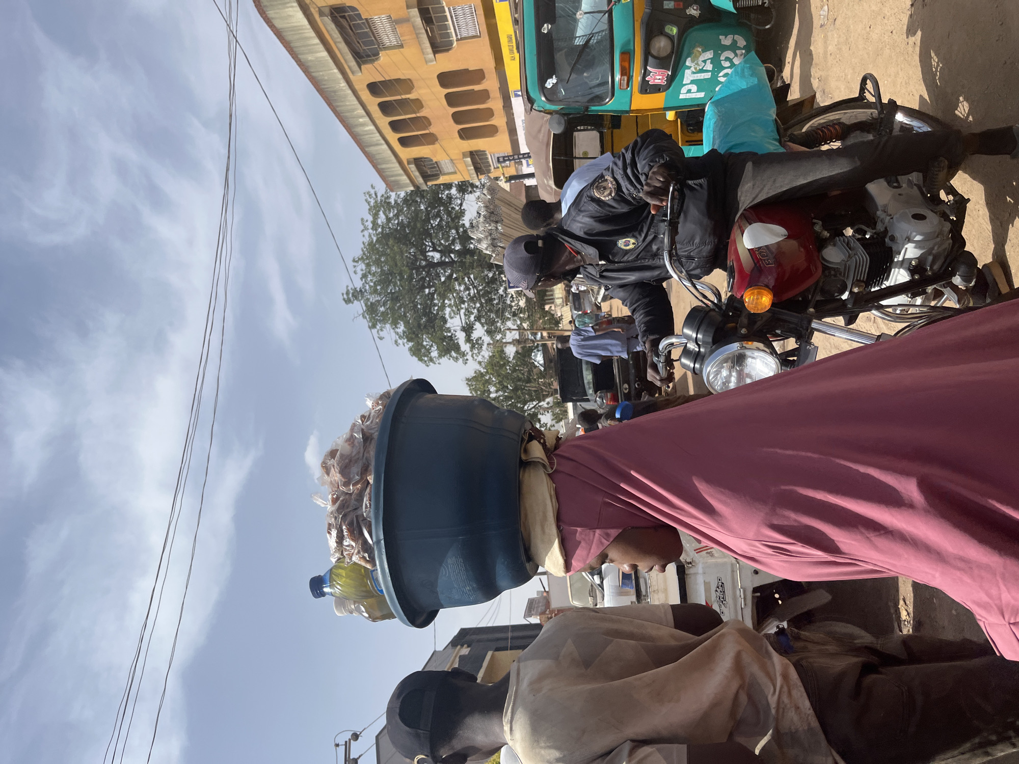 The image depicts a street scene in Jos, Nigeria. A woman is carrying a large blue basin filled with items, including what appears to be food in plastic bags and bottles of liquid. She is wearing a pink garment. In the background, a man is sitting on a red and silver motorcycle. Behind him, there is a green and yellow auto-rickshaw. Other people are visible in the middle ground. Buildings are visible in the background. The sky is partly cloudy. Power lines are visible in the upper left of the frame.