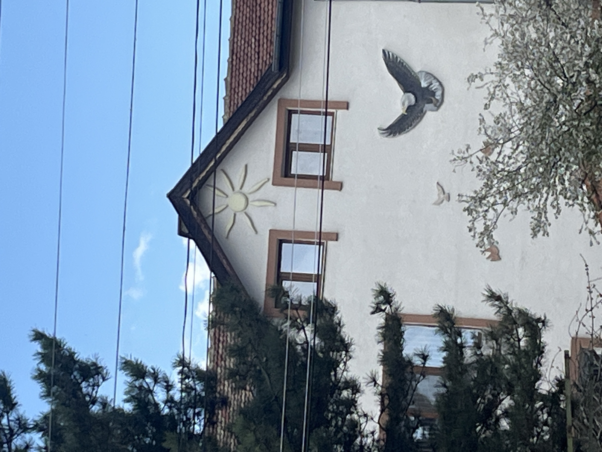 A partial view of a multi-story building with a white facade, terracotta tiled roof, and brown-framed windows is depicted. The building features decorative elements, including a sun symbol on the gable and bird sculptures in flight on the facade. A tree with sparse foliage partially obscures the right side of the building. Pine trees are visible in the foreground. Overhead power lines intersect the sky. The sky is blue with scattered clouds. The location may be in Burgas, Bulgaria.