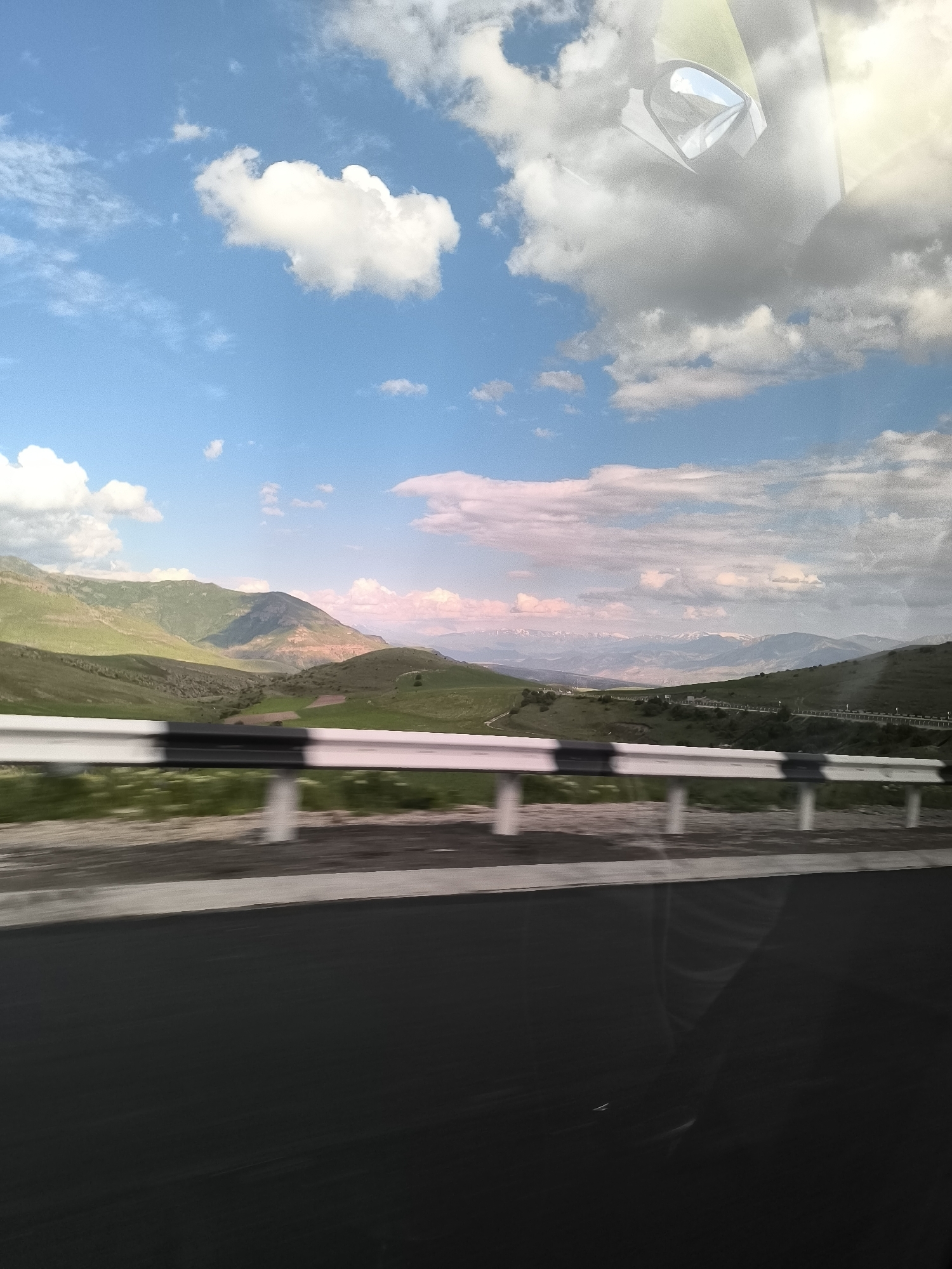 The image captures a panoramic view of the mountainous landscape of Yelpin, Armenia, taken from a moving vehicle. The scene is dominated by a vast blue sky, dotted with fluffy white cumulus clouds and larger grey cloud formations. The light appears soft, possibly late afternoon, with subtle pink and golden hues on the distant clouds and mountains.
In the mid-ground, rolling green hills and mountains stretch across the horizon, some displaying darker, rugged patches. Further in the distance, snow-capped peaks are faintly visible, adding to the grandeur of the natural setting.
The foreground features a black asphalt road, suggesting a highway or a well-maintained route. A white and black striped guardrail runs along the side of the road, exhibiting motion blur, which indicates the photograph was taken while traveling at speed.
A notable detail is the clear reflection in the upper right quadrant of the image. It shows parts of a vehicle's interior, specifically a side mirror and what appears to be elements of the car's ceiling or windshield, confirming the perspective from inside a moving car. This reflection suggests the photograph was taken through a window.
The overall impression is one of a scenic journey through a picturesque, mountainous region under partly cloudy skies. There is no visible text in the image.