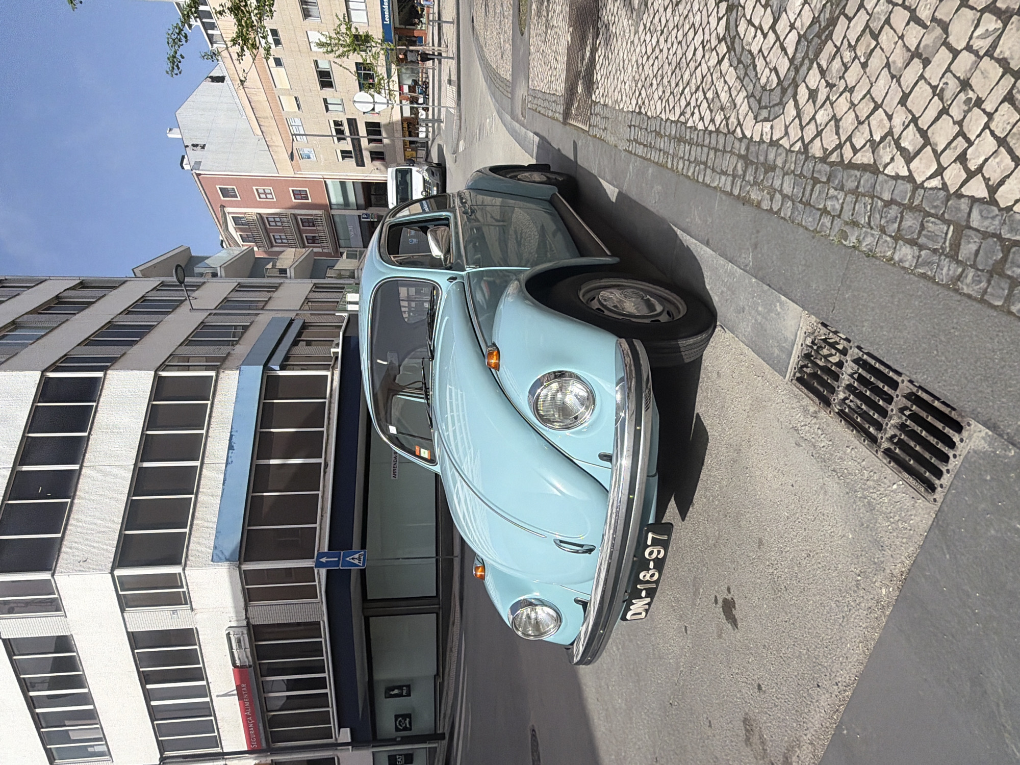 A light blue Volkswagen Beetle is parked on a street in Aveiro, Portugal. The car's license plate reads "DN-18-97". The location appears to be a commercial area, with tall buildings, shops, and cobblestone sidewalks. A building with numerous windows is visible on the left side of the car. A street sign with a left-pointing arrow is mounted on the building. There is a storm drain next to the car. Other buildings are in the background. The weather seems sunny.