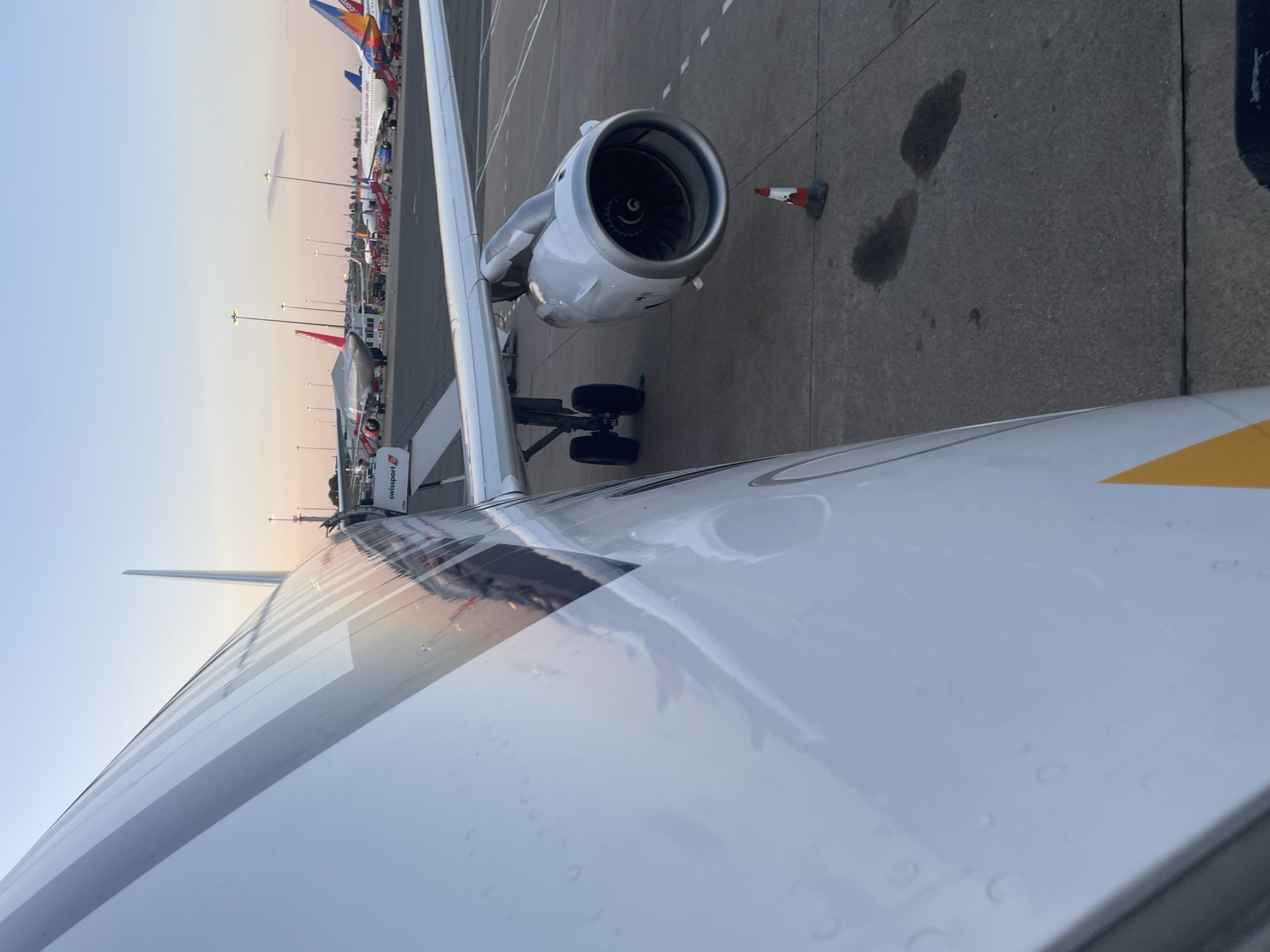 The image shows a close-up view of an airplane's engine and wing from an adjacent aircraft, likely at Birmingham Airport in the Metropolitan Borough of Solihull, United Kingdom. The perspective is from inside an aircraft, looking out at the tarmac. Several other aircraft are visible in the background, parked at the gate or on the runway, with a clear sky and setting sun casting a warm glow. The tarmac area shows a traffic cone, some dark fluid stains, and other markings. The aircraft in the foreground displays the "swissport" logo on a visible boarding stair. The time of day appears to be dusk or early evening.