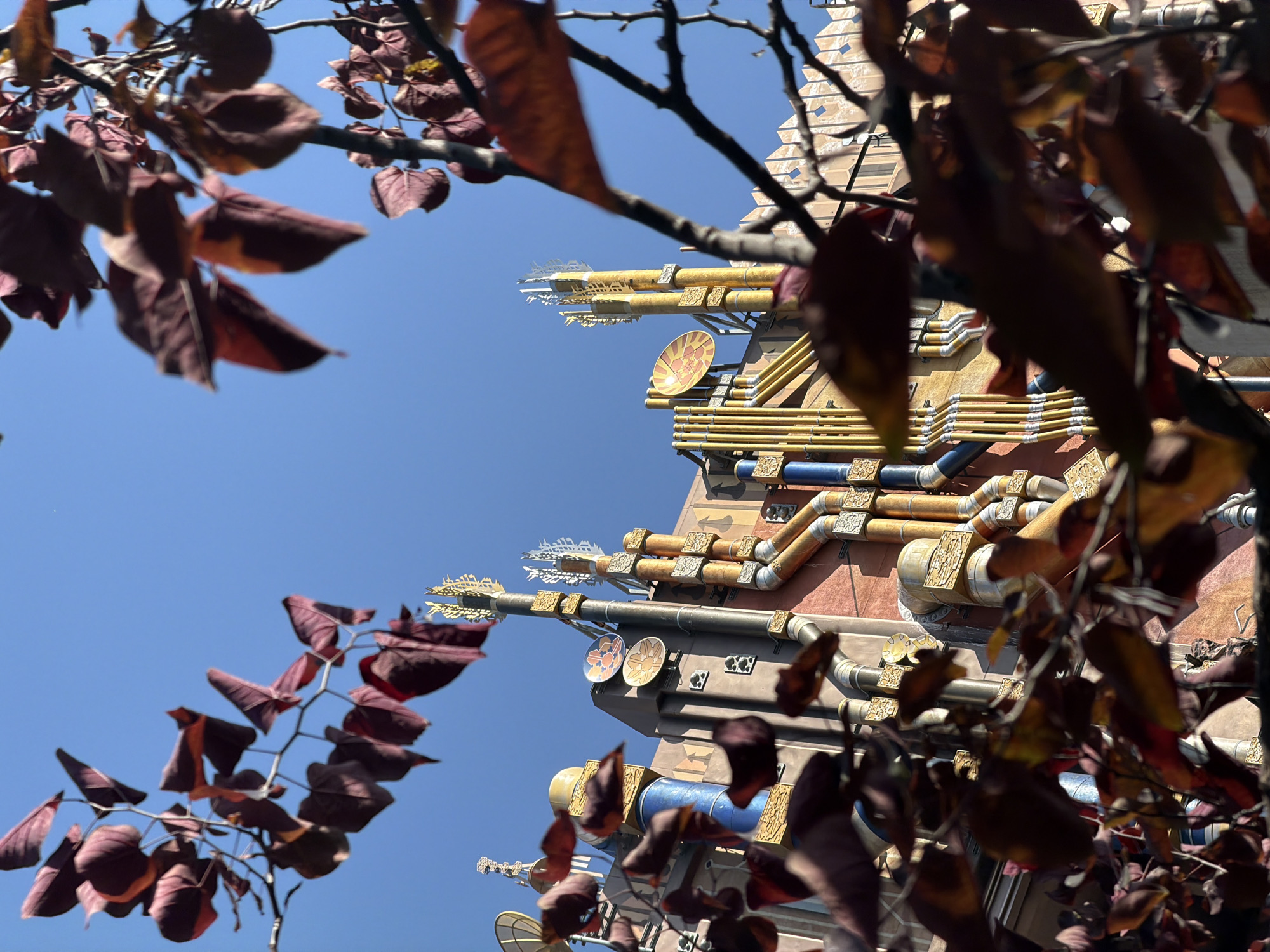 The photograph, taken from a low angle, shows a building structure with ornate, possibly steampunk-inspired, architectural details against a clear blue sky. The building's exterior is embellished with various metallic fixtures, including cylindrical pipes, decorative plates, and intricate spires. The color palette of the building includes gold, silver, and copper tones. In the foreground, tree branches with red leaves partially obscure the view of the structure.
Based on the architectural style and details, the building is likely located in the Star Wars: Galaxy's Edge area of Disneyland in Anaheim, California, United States of America. The scene depicts elements of science fiction and fantasy, creating an immersive environment.