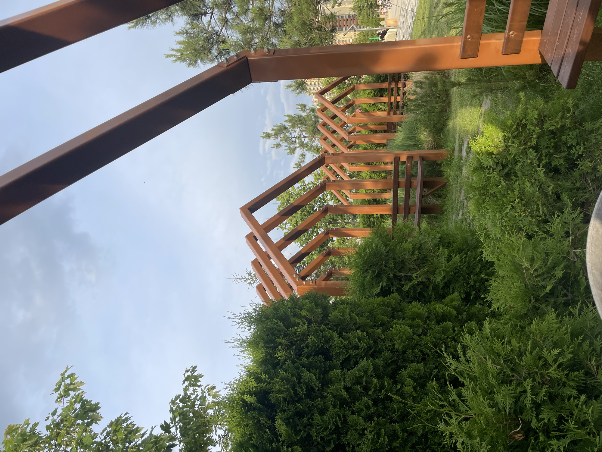The image depicts a park area with several wooden pergolas forming a repetitive pattern of arched structures. Each pergola is designed with a wooden frame that suggests a roof shape and is supported by vertical beams. Benches are placed within and around these structures, some occupied by unseen individuals. Lush green foliage, including a large, dense bush in the foreground and other ornamental plants and trees, surrounds the pergolas. The ground is covered with grass and paved pathways. In the background, a modern building complex is visible, along with more trees and a cloudy sky. The scene is likely located in the "Dolina Promyslov" park in Stavropol, Russia, given the architectural style of the pergolas which are a common feature of the park's recent renovation.