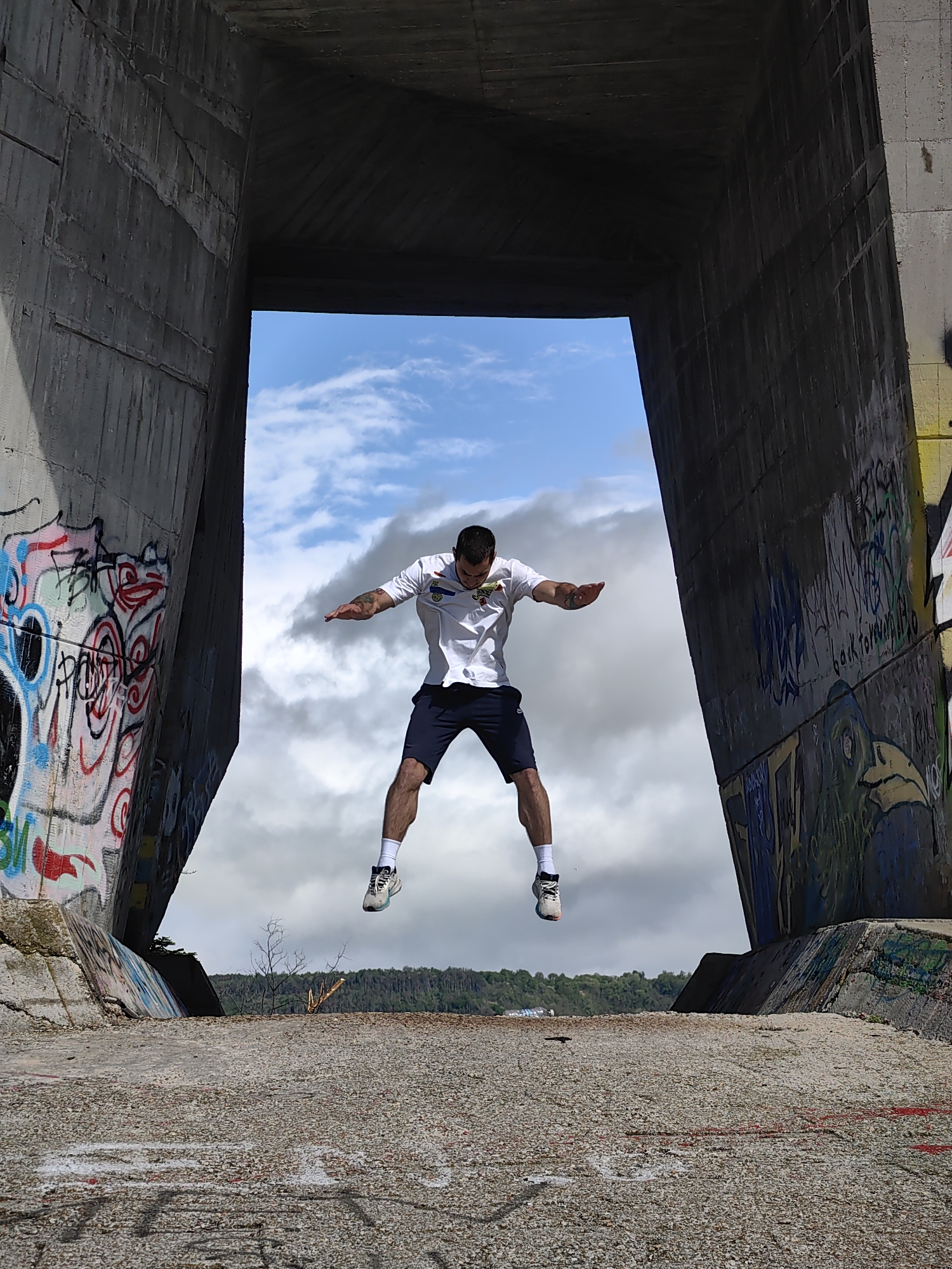 A man is suspended in mid-air within a concrete structure. The structure is adorned with graffiti on its sides. A sliver of green hillside is visible through the structure, topped by a sky filled with white clouds. The man is light-skinned, wearing a white shirt, dark shorts, white socks, and running shoes. The ground, a concrete surface, displays more graffiti. The scene is at the Asparuhov Bridge in Varna, Bulgaria.