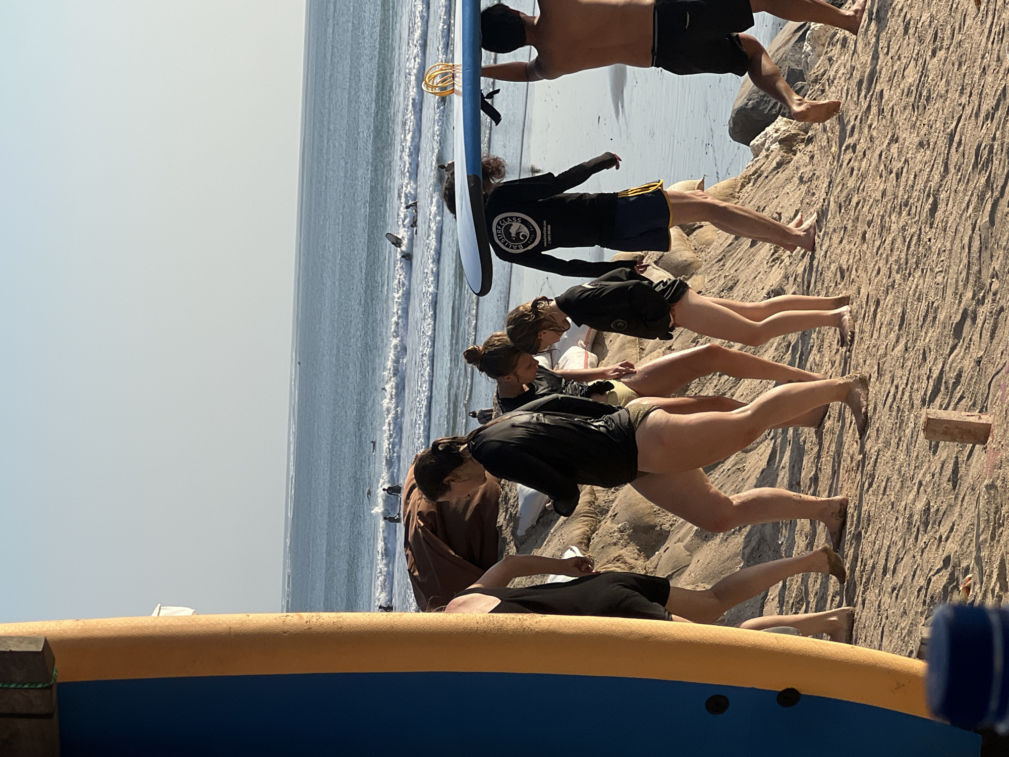 The image depicts a group of people, likely a surf class, practicing on a sandy beach near the ocean. Several individuals are positioned on the sand, appearing to be engaged in a physical activity, possibly a warm-up or a specific exercise related to surfing. Their bodies are oriented in various poses, with some in a prone position on their stomachs and others in a more upright stance.
The scene is set on a beach with the ocean in the background. Small waves are visible breaking near the shore, suggesting it's a coastal environment suitable for surfing. The sand appears dry and coarse, with some rocky formations present. The lighting suggests it is daytime, with the sun casting shadows and illuminating the scene. The weather appears clear and sunny.
One individual in the foreground is wearing a black long-sleeved rash guard with yellow trim on the shorts. Another person is wearing a similar black rash guard and dark shorts. The attire is typical for surfing or water sports.
On the back of the black rash guard of one individual, there is visible text. It appears to be a logo and some writing, possibly the name of the surf school or event. While not entirely legible due to the resolution and angle, phrases like "SURF CLASS" and potentially an organization name can be discerned.
The overall mood of the image seems to be active and focused, with the participants concentrating on their movements. The presence of surfboards, including a blue and white one in the upper part of the image, further confirms the surfing context. The location is Kuta, Indonesia, which is a well-known surf destination.