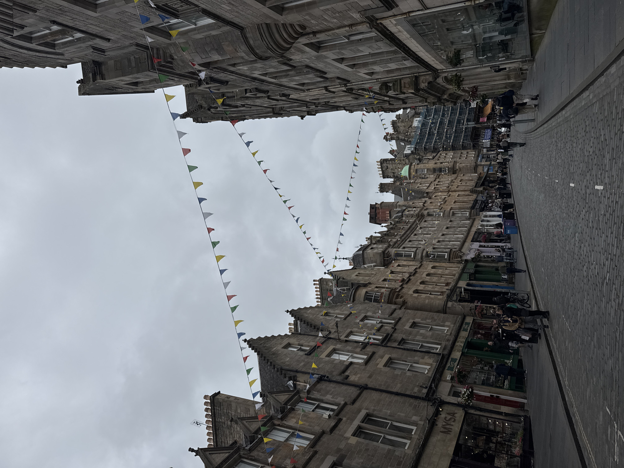 A street scene in Edinburgh, United Kingdom, features a cobblestone road lined with multi-story stone buildings. Strung between the buildings are triangular flags in various colors, including red, yellow, green, and blue. The sky is overcast and gray. Several people are visible walking along the sidewalks and crossing the street. The buildings have traditional stone facades with numerous windows and decorative architectural elements. Some of the ground-floor businesses have awnings and shop windows displaying goods. Scaffolding is present on one of the buildings further down the street, indicating ongoing construction or renovation.