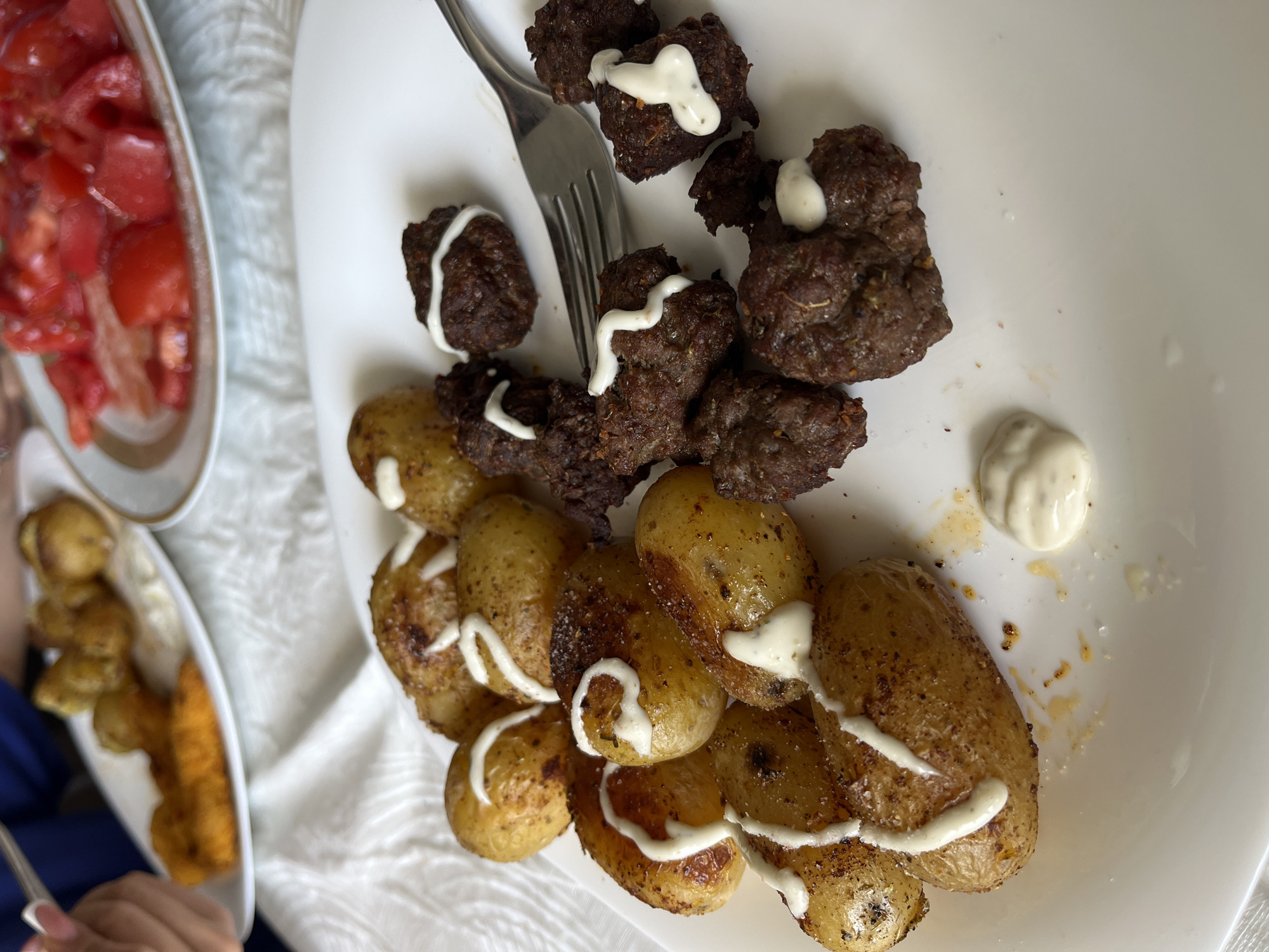 On a white plate, small fried potatoes are arranged next to small, dark, round meat patties. Both the potatoes and meat patties are topped with a white sauce. A fork pierces one of the meat patties. To the left, out of focus, is another plate of red tomatoes and fried food. The photo was taken in the city of Zlatograd, Bulgaria.