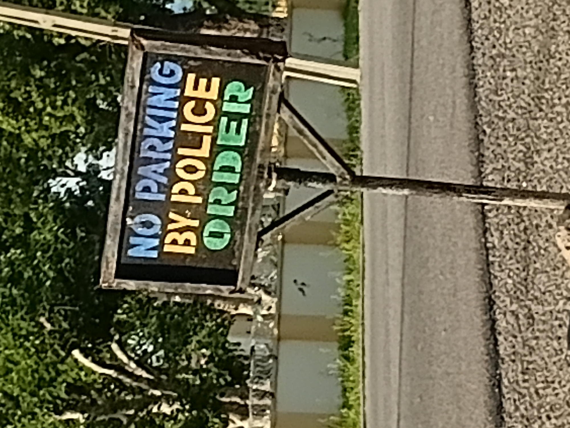 A vintage "NO PARKING BY POLICE ORDER" sign is mounted on a wooden post. The sign features blue lettering for "NO PARKING" and yellow for "BY POLICE," with green lettering for "ORDER." The sign itself appears weathered, suggesting it has been in place for some time. It is situated in an outdoor environment with lush green trees visible in the background. A light-colored, possibly concrete or stucco, wall with some decorative elements runs horizontally behind the sign. The foreground consists of a gravelly area adjacent to a paved road. The scene is set in Jalingo, Nigeria. There are no people or animals prominently visible in the frame.