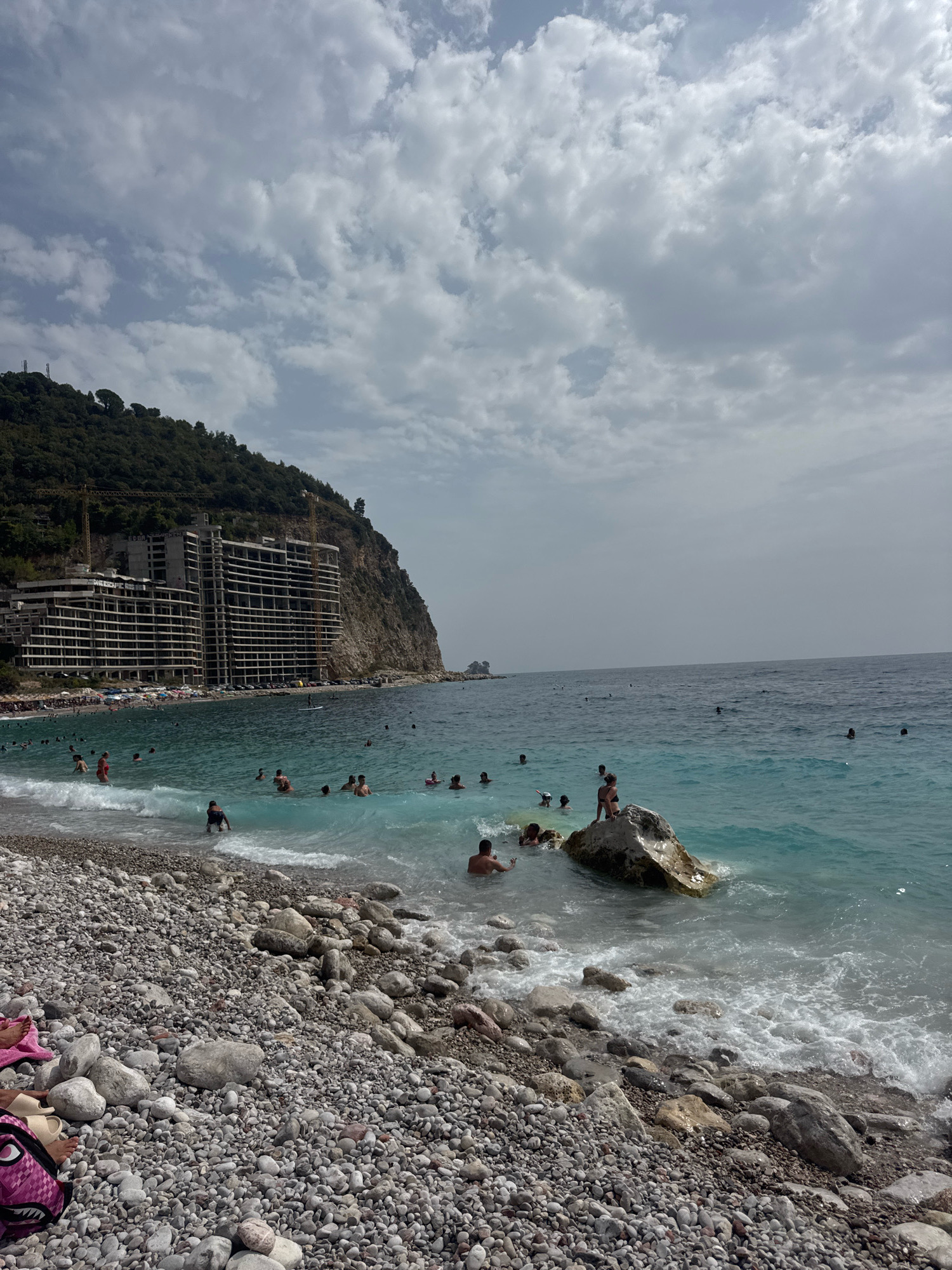 This is a wide-angle, eye-level shot of a crowded, pebble beach with turquoise waters in Mirište, Montenegro. In the background, a massive, multi-story building, possibly under construction, dominates the hillside. Two yellow construction cranes are visible on the left side of the building. The sea is calm with gentle waves lapping onto the shore. Many people are swimming in the water, some are standing on a large rock near the shore, and others are scattered on the beach among the pebbles and beach towels. The sky is cloudy with patches of blue visible.
