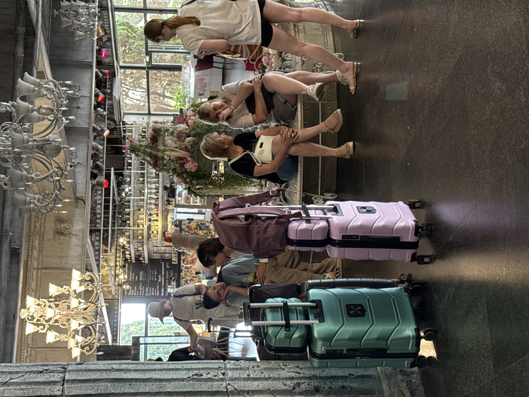 In the city of Yalta, Ukraine, within what appears to be a café or hotel lobby, two teal and lavender suitcases stand prominently in the foreground. The teal suitcase is a larger, lower model, while the lavender one is a smaller, stacked unit. To the right of these, a person stands facing away from the camera, wearing a white t-shirt and black shorts. In the middle ground, a group of four individuals are seated on a bench. Two women are engaged in conversation, one with blonde hair wearing a black t-shirt and shorts, and the other with blonde hair wearing a white t-shirt and gray shorts. A younger person with brown hair is also seated, looking towards the left. Further back, a woman in a white hat and t-shirt is visible near a counter with shelves stocked with bottles. The environment features decorative chandeliers hanging from the ceiling and a floral arrangement. The flooring is a dark, speckled material.