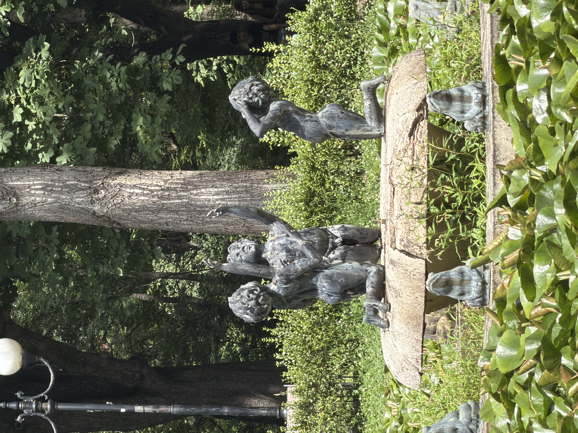 In Sofia, Bulgaria, within the South Park, a bronze fountain depicts three cherubic figures. Two are kneeling on a round, textured surface, one with its arms raised to the sky and the other with its head bowed. A third figure kneels to the right, clutching its head. They are positioned within a pond filled with lily pads. Surrounding the pond is manicured green shrubbery. In the background, large trees with rough bark form a dense canopy, and a vintage lamppost stands to the left. Two individuals are seated in the far background, partially obscured by foliage. The lighting suggests it is daytime, with strong sunlight casting shadows.