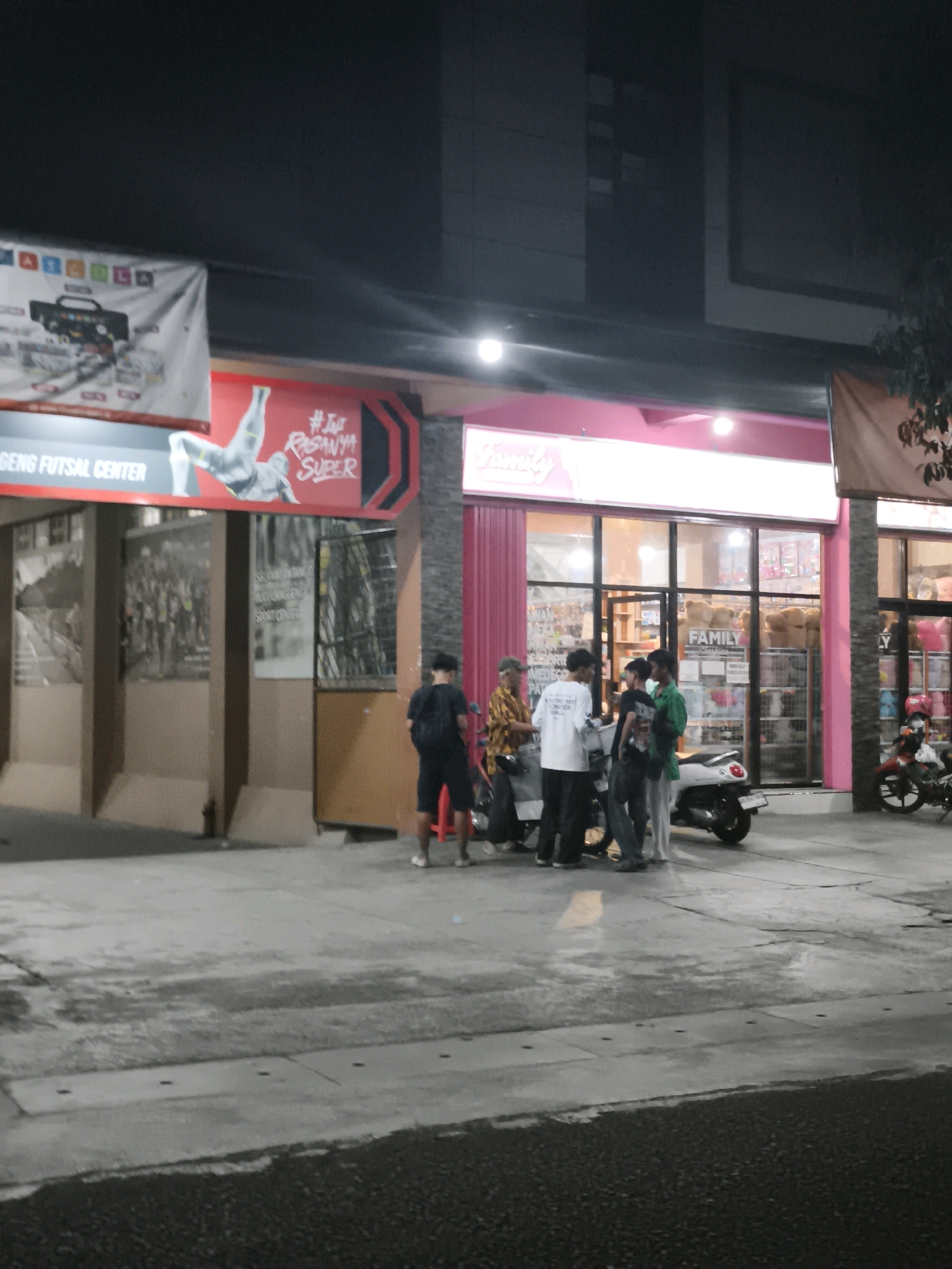 The image shows a nighttime street scene in front of a store. There are four men standing near a scooter, and another man is standing further to the right. The store appears to be a "Family" branded shop, possibly a convenience store or supermarket, with its interior lit and visible through large windows. In the background, a banner for "GENG FUTSAL CENTER" is visible. The pavement is wet, suggesting recent rain. The overall atmosphere is urban and commercial. It is located in Desa Sawahan, Indonesia.