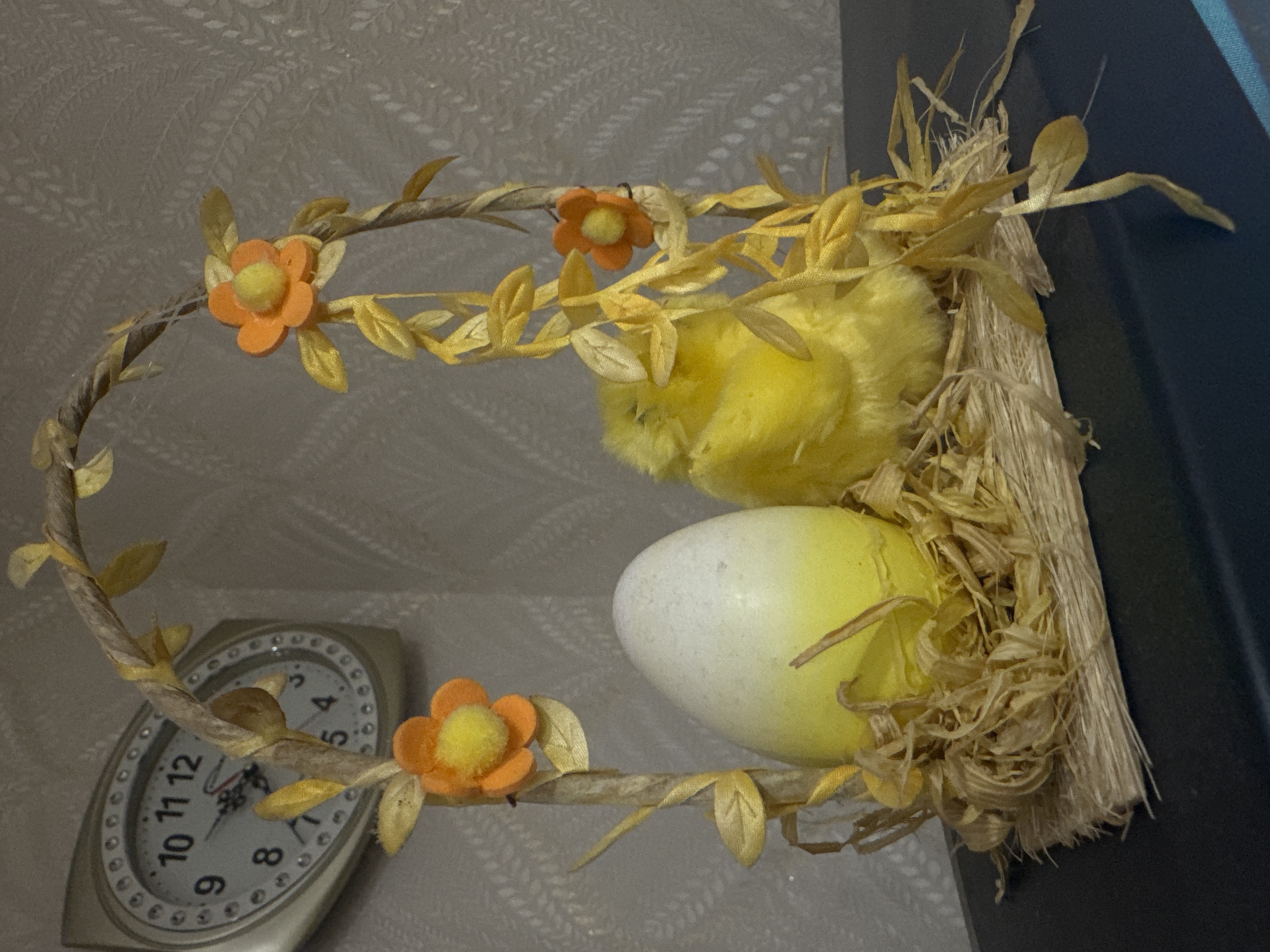 The image displays a decorative, spring-themed arrangement, likely an Easter decoration. The central subject is an oval-shaped arch, seemingly crafted from twisted natural fibers, adorned with small, stylized yellow leaves and three orange fabric flowers, each with a fluffy yellow center.
Nestled within the upper right curve of the arch is a bright yellow, fluffy chick, partially obscured. Below it, a large, ovoid egg rests in a bed of light brown, straw-like material, forming a nest. The egg features a gradient color, transitioning from white on one end to light yellow on the other, with subtle speckles across its surface. The straw nest is substantial and extends beyond the arch on the right side.
In the background, on the lower left side, a silver-toned, oval-shaped analog clock is visible. It has a white face with clear black Arabic numerals from 1 to 12. The hour hand points just past 9, and the minute hand is on the 4, indicating the time is approximately 9:20. A thin red second hand is also present. The clock's bezel is decorated with small, circular elements. The wall behind the decoration has a subtle, light-colored patterned texture. The overall composition suggests an indoor setting with a festive, seasonal theme. No specific activity is depicted, as it's a still life arrangement.