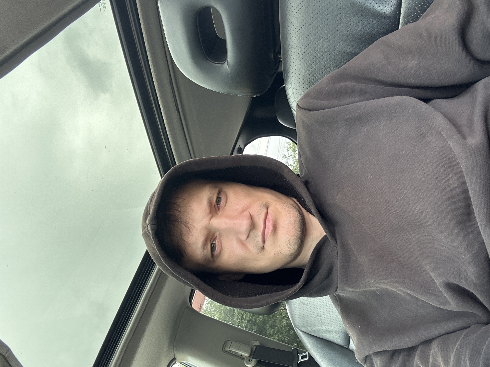 A man wearing a dark brown hooded sweatshirt is sitting in the driver's seat of a car, looking at the camera. The car has a sunroof that is open, revealing a cloudy sky. The interior of the car is visible, including black leather seats. Outside the car, trees are visible. The man's expression is neutral. The photo was taken in the city of Makiv, Ukraine.