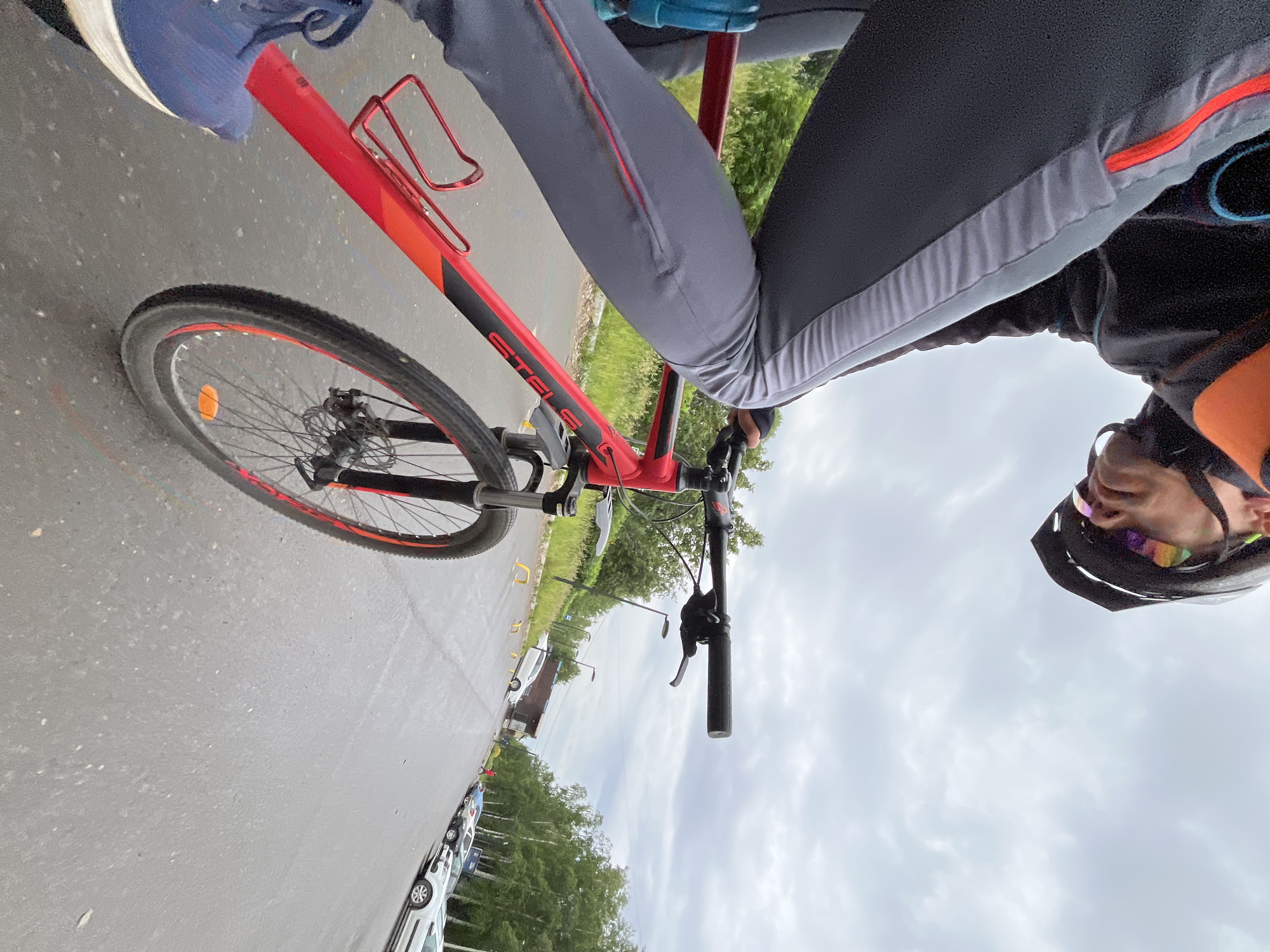 The image captures a first-person, high-angle perspective of a person riding a bicycle on a road in Chaikovsky, Russia, under an overcast sky.
The primary subject is a person, partially visible, wearing a black cycling helmet with colorful, iridescent lenses on their glasses, a dark long-sleeved top with orange accents, and grey cycling pants featuring red and light grey stripes. Their hands are on the black handlebars of a red and black bicycle.
The bicycle, a "STELS" brand, is prominently featured. It has a red frame with black accents and a red water bottle cage mounted on the frame. The front wheel has a black rim with red highlights, a black tire, and an orange reflector on its spokes, along with a disc brake. A small blue object is visible near the seat post.
The setting is an outdoor road, likely asphalt, appearing somewhat damp. Alongside the road, there are rows of parked white cars and green trees and bushes lining the environment. The sky is entirely cloudy, indicating an overcast day. The overall impression is of a cycling journey or stop on a cloudy day.