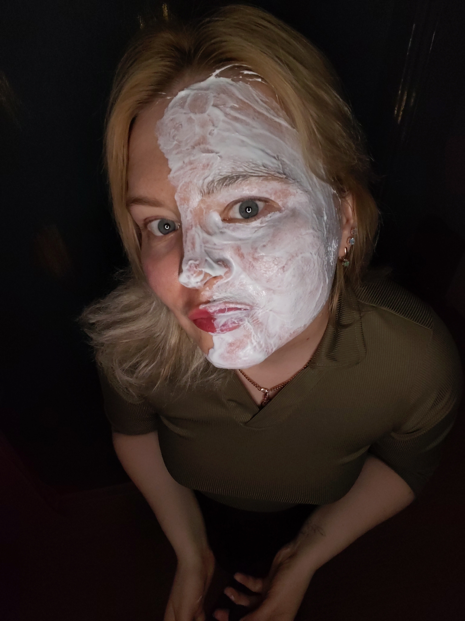 A woman with blonde hair is depicted in a low-angle, close-up shot. The right side of her face is covered in a thick, white substance, possibly a facial mask. Her left side of the face is uncovered and shows her bare skin with red lipstick. She has light eyes. She is wearing a green ribbed top and a gold necklace. Her hands are visible in the lower part of the frame. The background is dark and indistinct. There is nothing to indicate the location of Izhevsk, Russia.