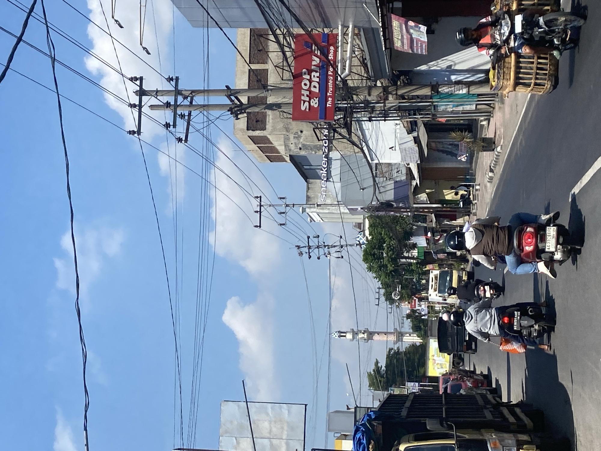 A street scene in the city of Kediri, Indonesia, depicts traffic on a sunny day. The foreground features several motorcycles with riders, some carrying passengers or cargo. One rider on the right is transporting large woven baskets on their vehicle. On the left, a large truck is visible. The street is lined with buildings and shops, including a prominent red sign that reads "SHOP & DRIVE The Trusted Parts Shop". A minaret of a mosque is visible in the distance, along with power lines and utility poles crisscrossing the sky, which is blue with scattered clouds.