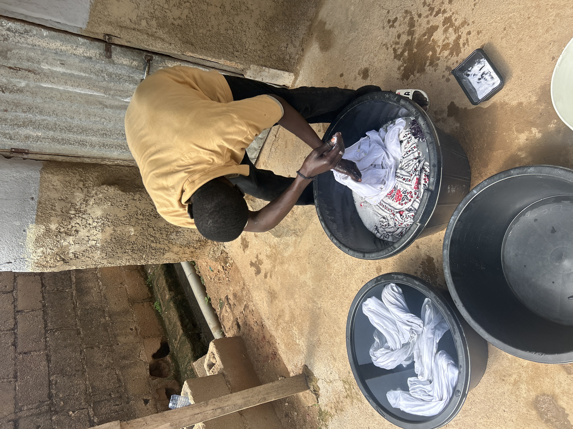 In a dusty outdoor courtyard in Jos, Nigeria, a young man wearing a mustard-colored t-shirt and black trousers is doing laundry. He is bent over a large black plastic basin, actively washing clothes by hand. White garments are submerged in the soapy water, and he appears to be scrubbing a piece of patterned fabric. Another identical black basin sits to his left, filled with clean white clothes. A third, empty basin is positioned in the foreground. The environment is rustic, with a rough-hewn brick wall on the left, a corrugated metal door on the right, and a concrete floor. There are scattered puddles of water on the ground, indicating the ongoing activity. The scene suggests a typical domestic chore taking place in a residential area.