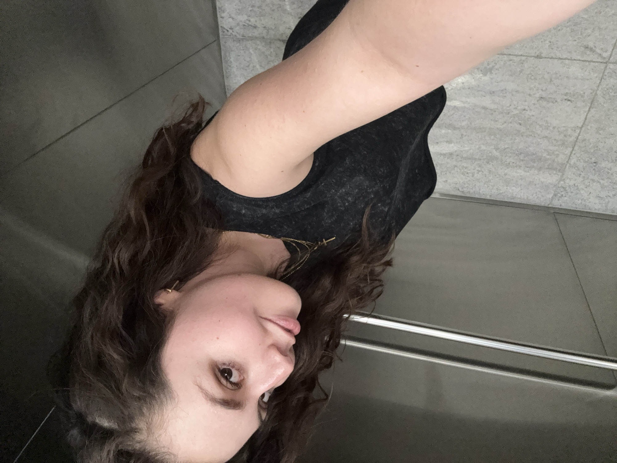 The image is a selfie of an adult female, rotated 90 degrees counter-clockwise. She has long, dark, curly hair, and fair skin, wearing a dark, textured top. Gold-toned necklaces are visible, one featuring a small cross pendant, along with a small gold earring in her right ear. Her expression is calm, and her gaze is directed towards the camera, appearing slightly upward and to the right within the frame.
The background indicates an indoor setting, possibly within a modern building in Mexico City, Mexico, such as an elevator or hallway. To the left, a smooth, dark-gray wall is visible, contrasting with the light-gray tiled flooring that occupies the right side of the image. A horizontal metallic bar extends across the lower right section, suggesting an architectural detail. The lighting appears to be artificial. There is no visible text in the image.