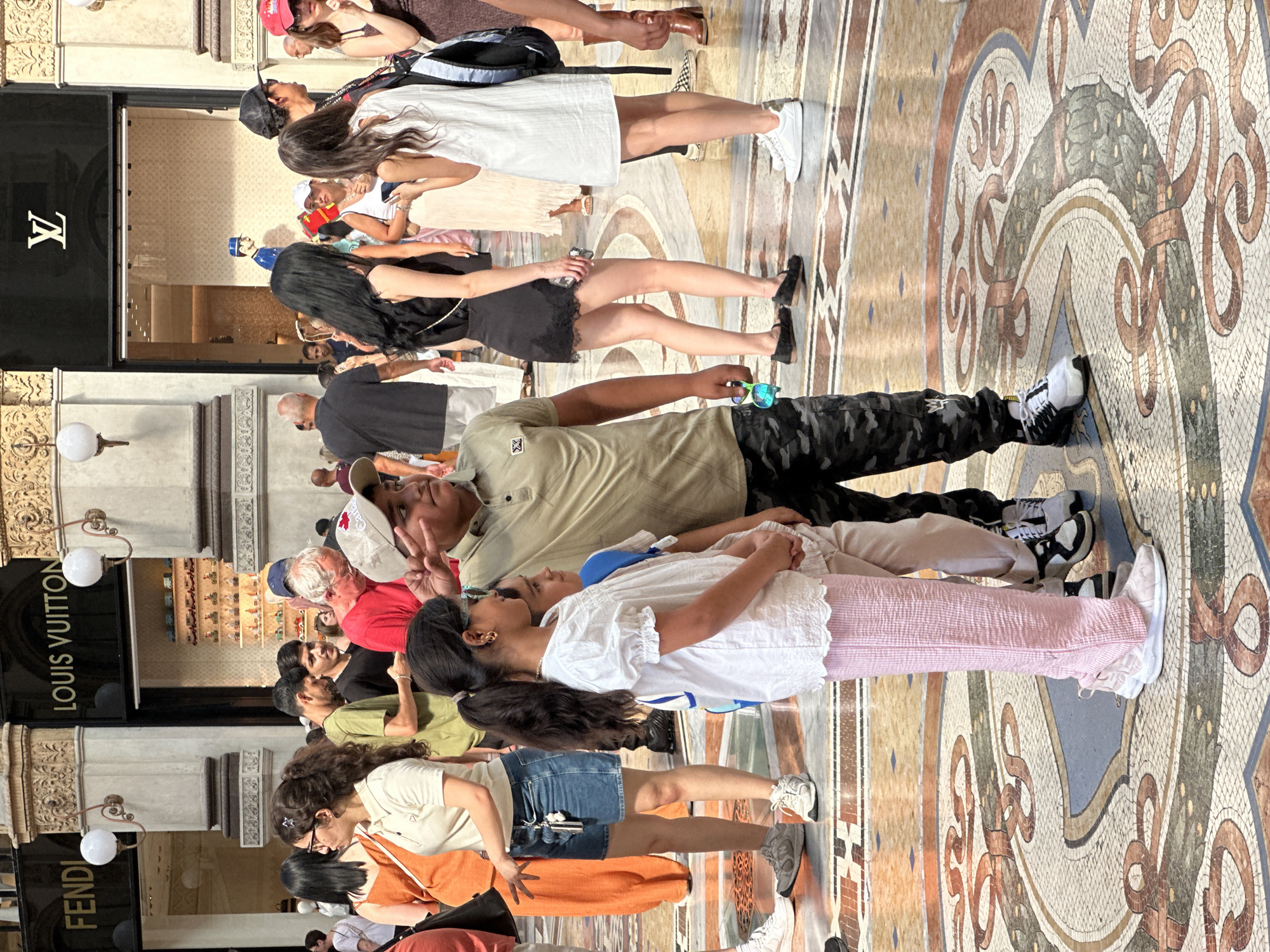 The image depicts a crowded interior scene, likely a high-end shopping arcade, with a mosaic floor featuring ornate designs. In the foreground, a young boy in a khaki shirt and camouflage pants stands at attention, saluting. To his left, a young girl in a white top and pink checkered pants looks to her side. Behind them, various individuals are visible, some browsing store displays or walking through the area. The background reveals storefronts with prominent branding, including "LOUIS VUITTON" and "FENDI," indicating a luxury retail environment. The architecture suggests an ornate and historic building, consistent with locations in Milan, Italy. The overall atmosphere is that of a busy public space with shoppers and onlookers.