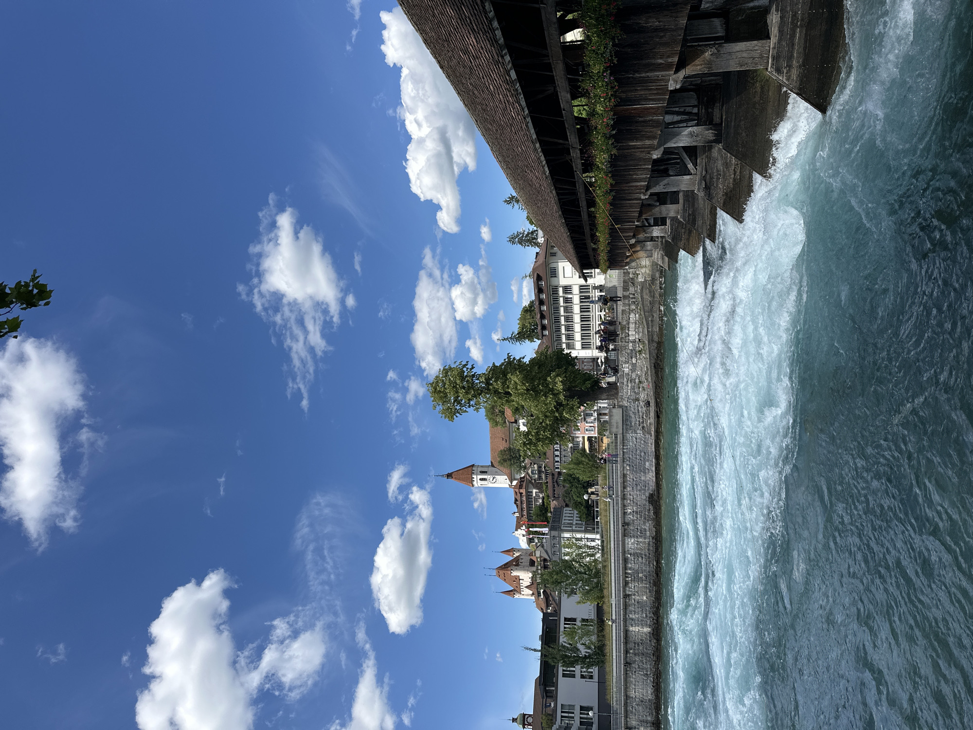 This is a photograph of the Aare river in Thun, Switzerland. The river flows rapidly in the foreground, with white water indicating strong currents. To the right, a historic wooden covered bridge spans the river, its weathered planks and roof visible. Flower boxes with red and green foliage adorn the sides of the bridge. In the background, the city of Thun rises, dominated by a large medieval castle with prominent towers and red-tiled roofs. A church with a tall, white steeple and a clock face is also visible. Buildings in the city appear to be a mix of modern and older architecture. A stone embankment runs along the riverbank, with a walkway on top where several people are casually strolling or standing. The sky is a vibrant blue, dotted with fluffy white clouds, suggesting a clear and sunny day. A tree branch with green leaves hangs into the frame from the top left corner.