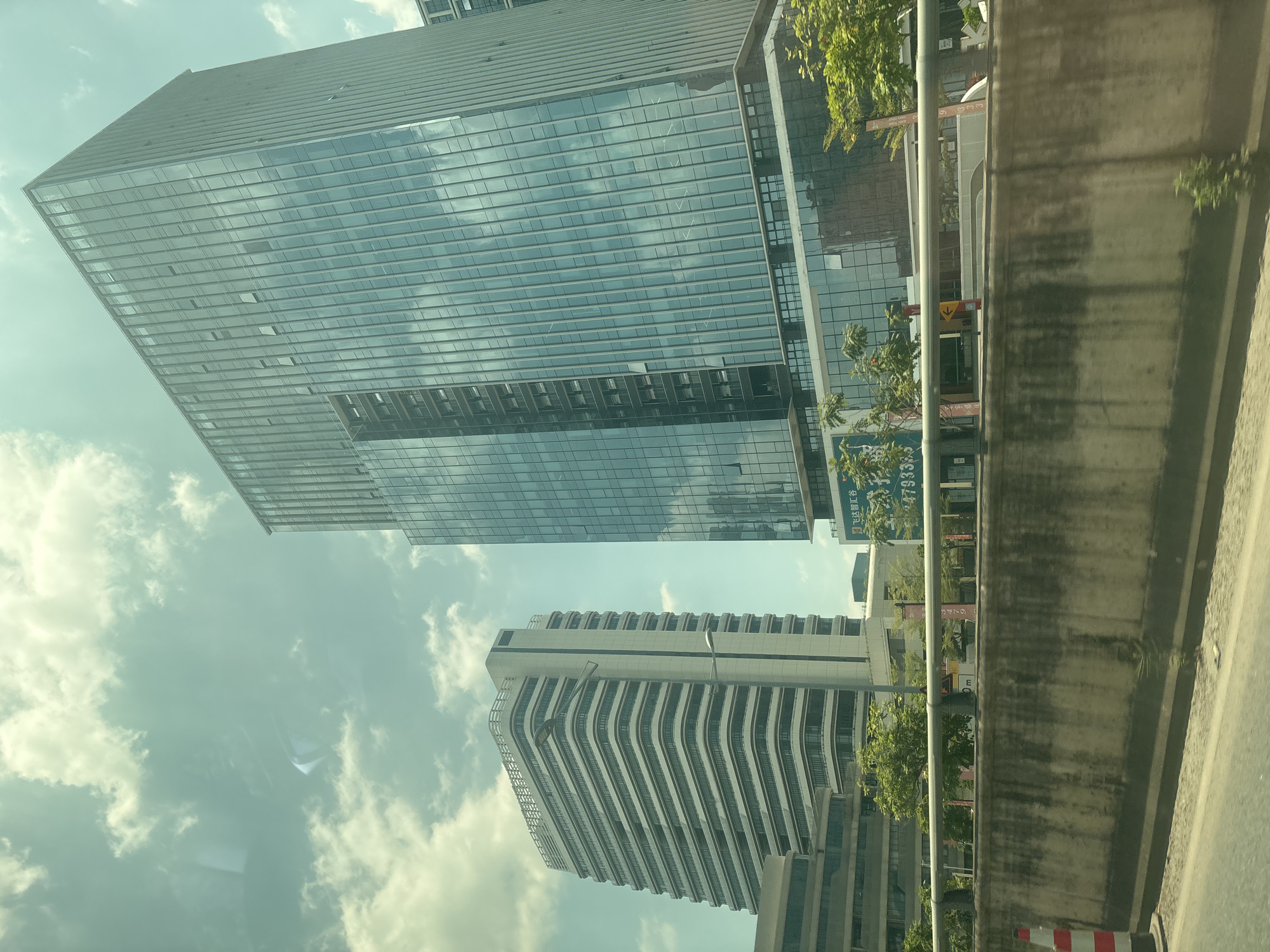 The image shows a view from a vehicle traveling along a road in Panyu District, China. In the background, two modern office buildings stand prominently against a sky with scattered clouds. The building on the right is a tall, sleek structure with a glass facade that reflects the sky and surrounding environment. The building on the left is also an office building, characterized by its white, layered exterior and horizontal window lines. A concrete barrier with a metal railing runs along the side of the road in the foreground. Trees and smaller buildings are visible between the road and the larger office structures. Signs with Chinese characters are affixed to the railing. The overall scene depicts an urban landscape with contemporary architecture.