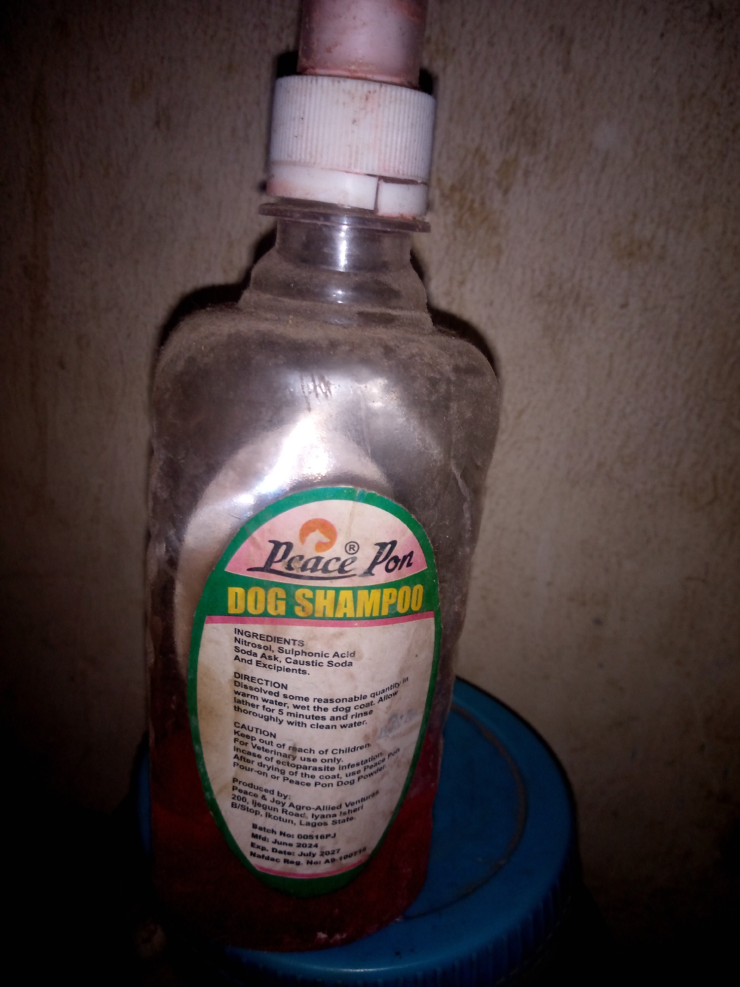 A close-up, low-angle shot shows a dusty plastic bottle of "Peace Pon Dog Shampoo" sitting on a blue plastic surface, possibly a lid, against a textured, dark wall. The bottle is partially filled with a red liquid, and the label is partially obscured by dust and the curve of the bottle. The label indicates the ingredients, directions for use, and a caution to keep out of reach of children. It also lists the producer as "Peace & Joy Agro-Allied Ventures" located at "200, Ijegun Road, Iyana Isheri B/Stop, Ikotun, Lagos State." The batch number is 00516PJ, manufactured in June 2024, and expiring in July 2027. The NAFDAC registration number is A9-100718. The lighting is dim and appears to be artificial, creating shadows and highlighting the dust on the bottle. There are no people or animals visible in the image. The environment is indoors, with a plain, slightly stained wall serving as the background. The location is not Jalingo, Nigeria, but Lagos State, Nigeria, based on the label information.